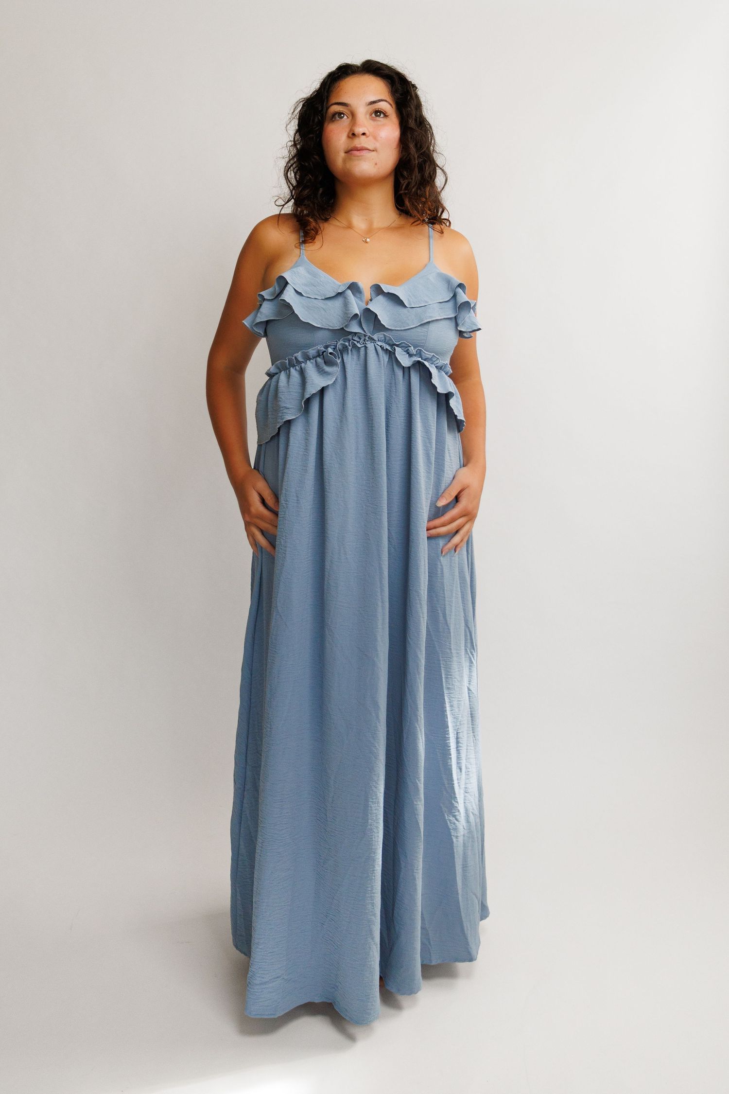 Pregnant woman wearing a dusty blue ruffle maxi dress with spaghetti straps, posing against a white background.