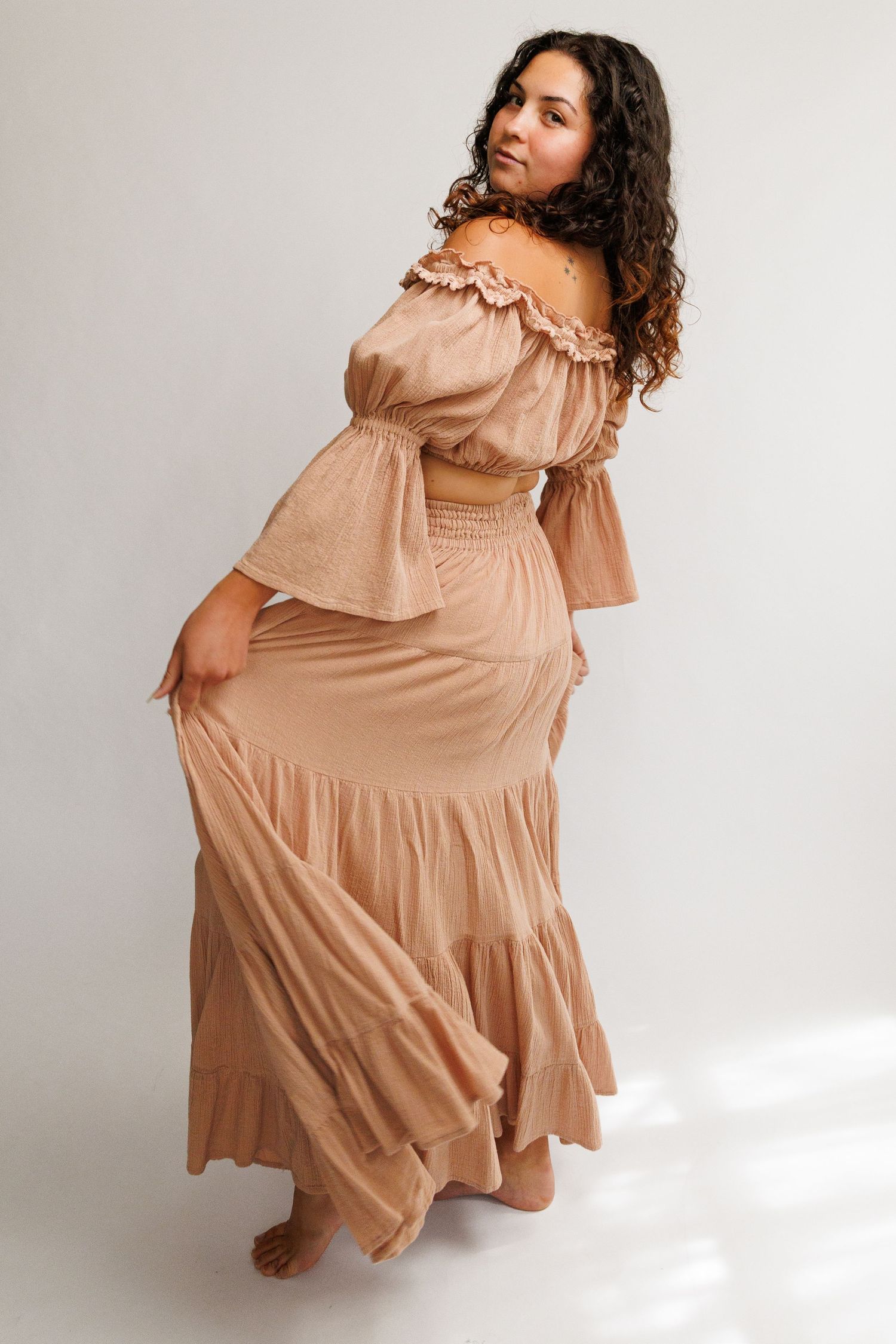 Woman wearing a flowing off-shoulder tiered maxi dress in warm beige, posing gracefully against a white background.