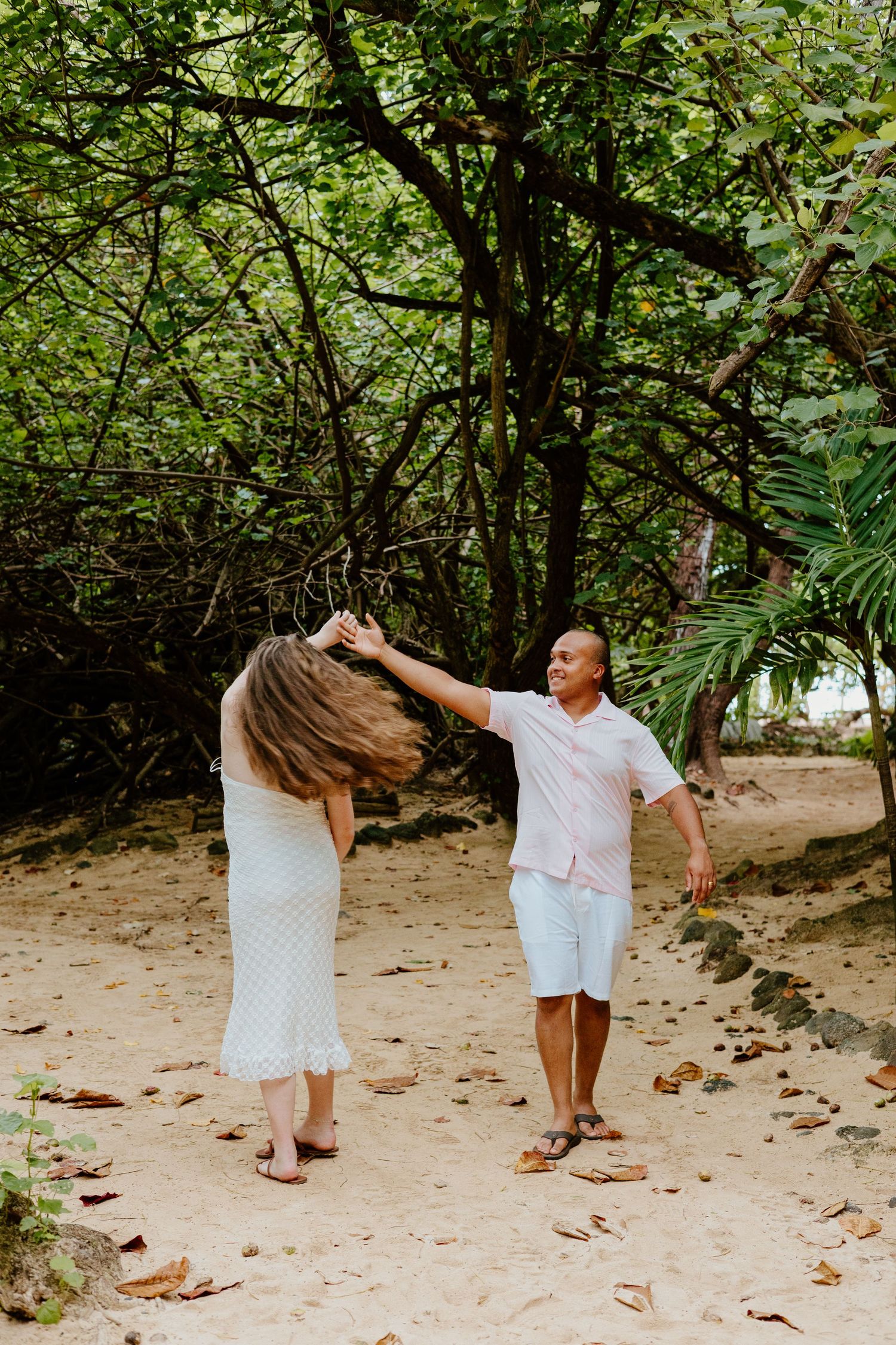 Couple dancing playfully on a sandy beach path surrounded by lush tropical trees and foliage.