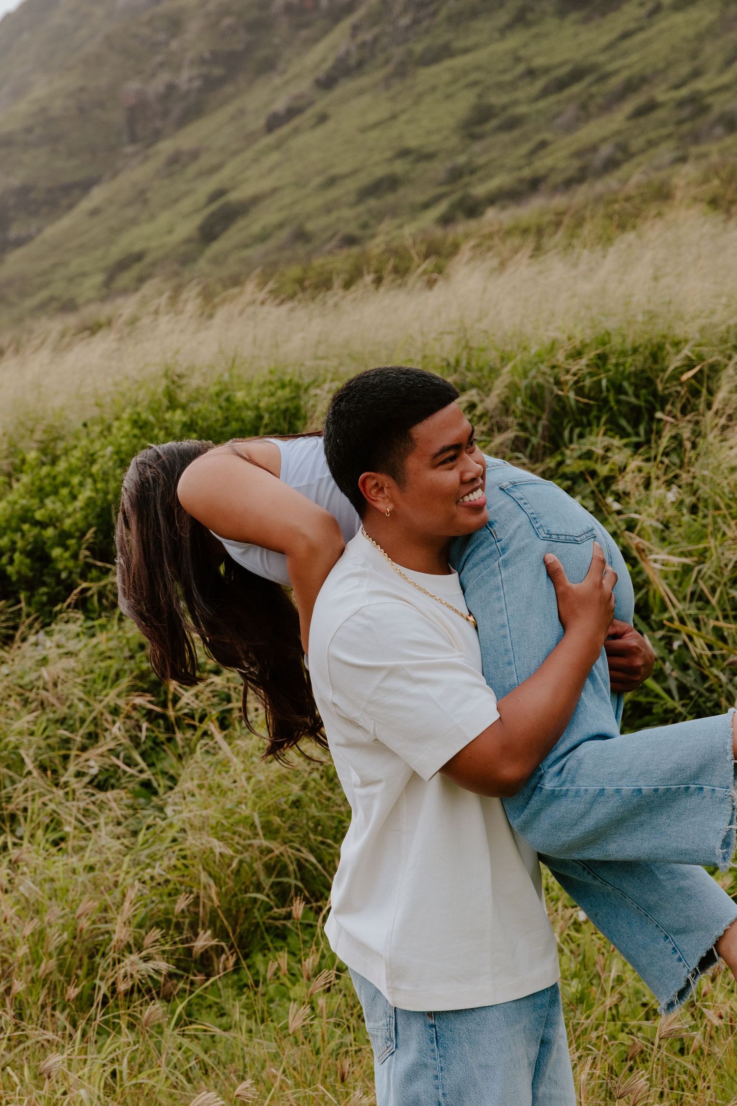 A smiling man carries a laughing woman on his back in a lush green Hawaiian hillside setting.
