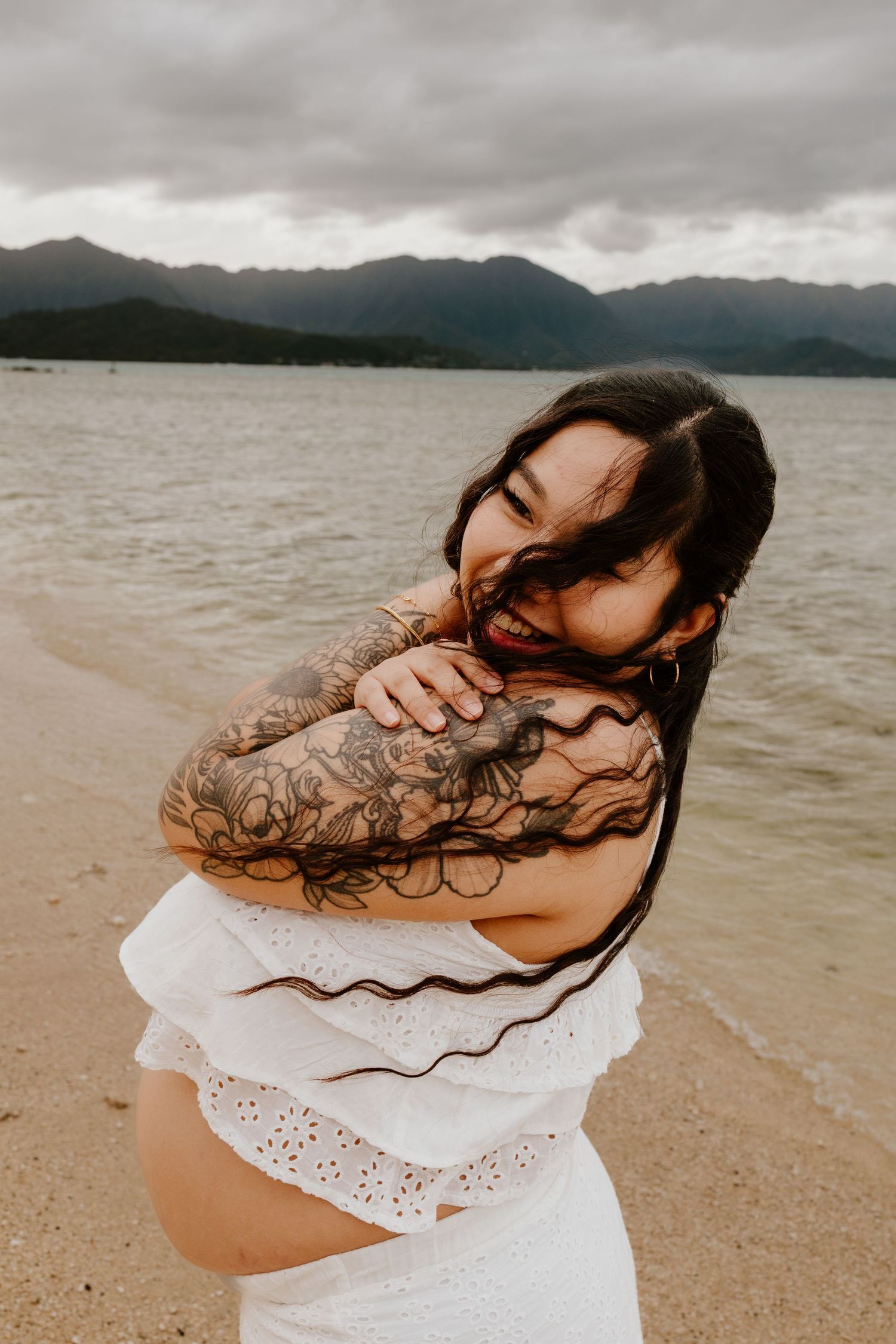 Smiling pregnant woman with tattoos in white dress embracing herself on a beach with mountains in background.
