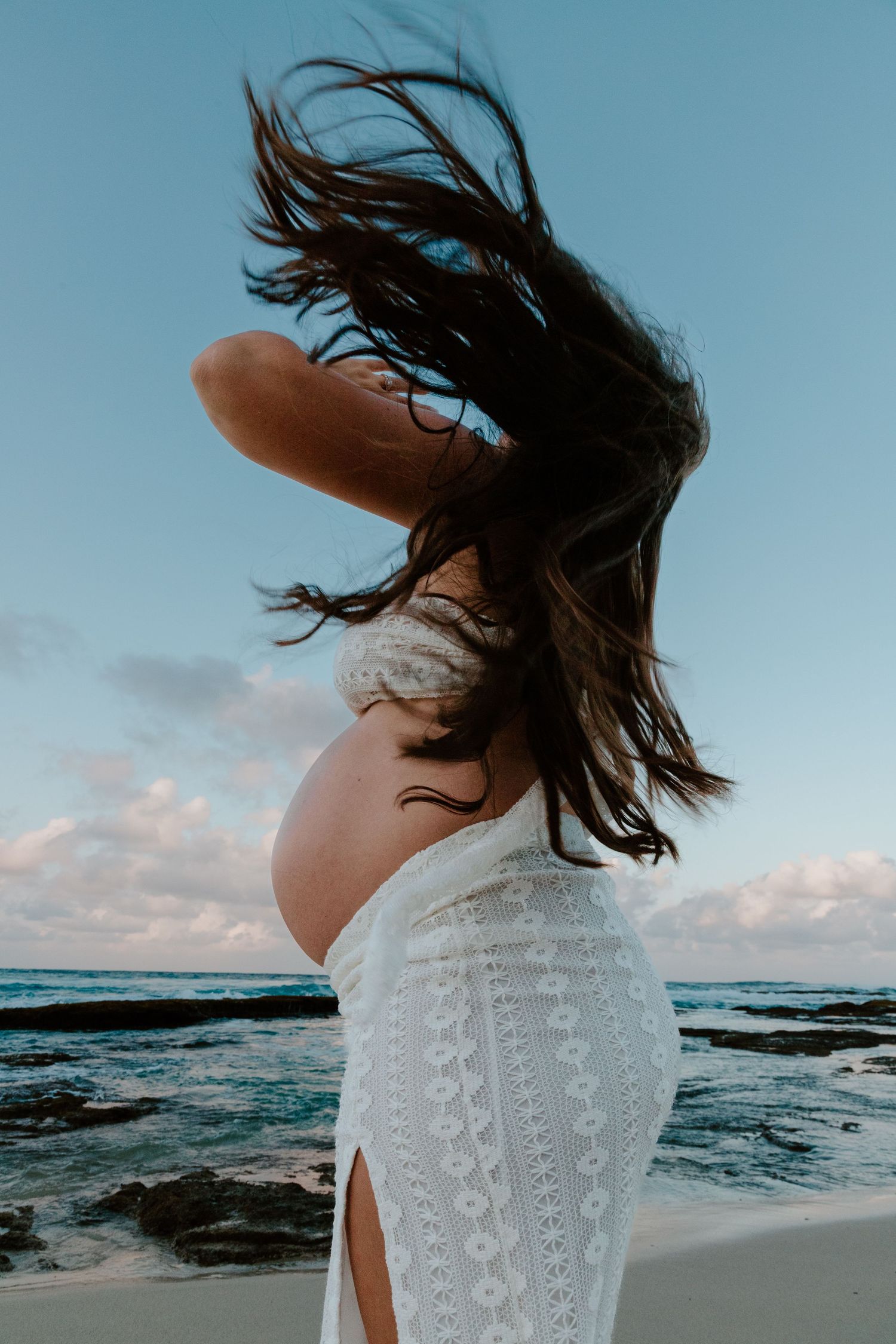 Pregnant woman tossing long dark hair on windy beach at dusk.
