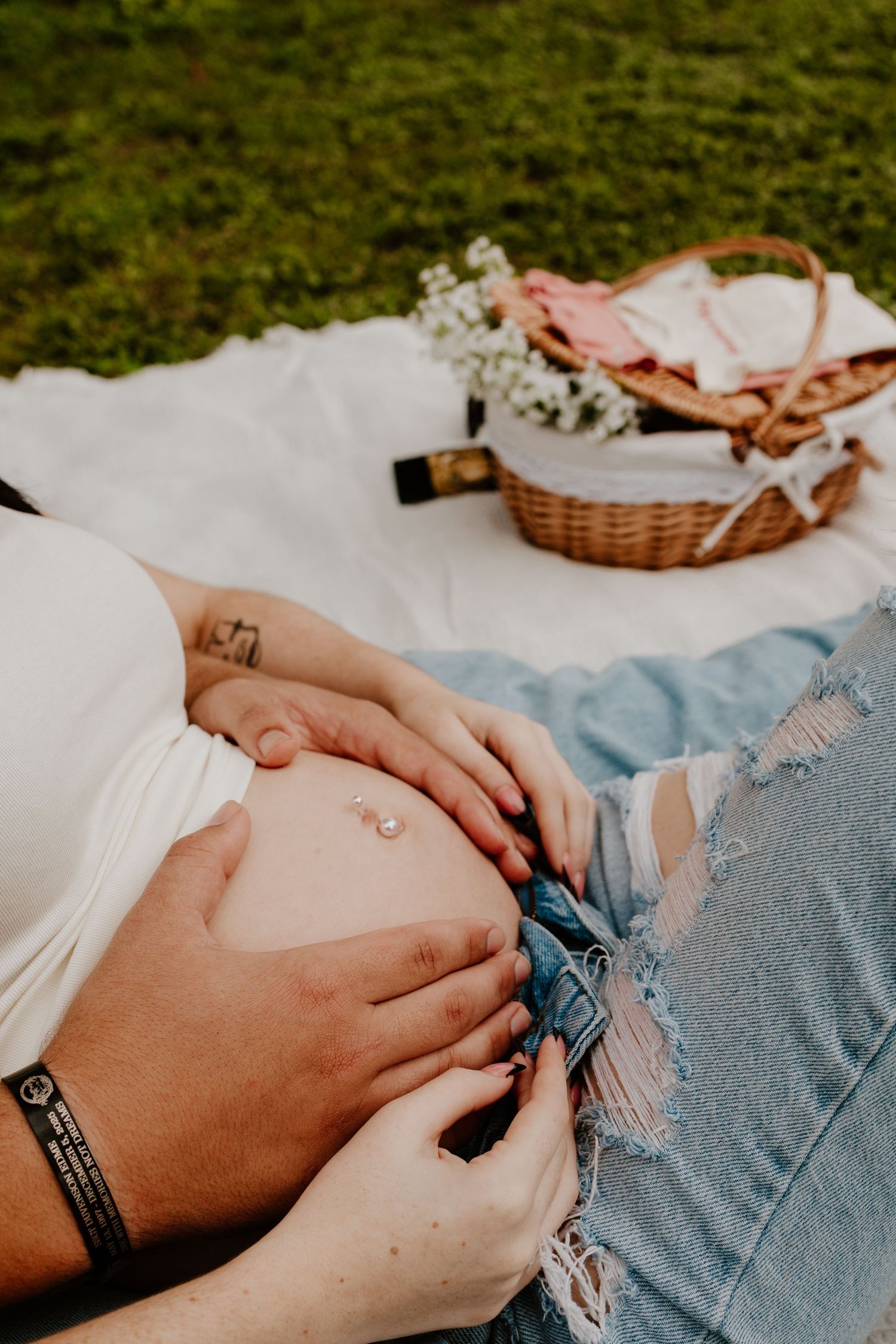 Close-up of pregnant belly on picnic blanket with wicker basket and white flowers nearby.