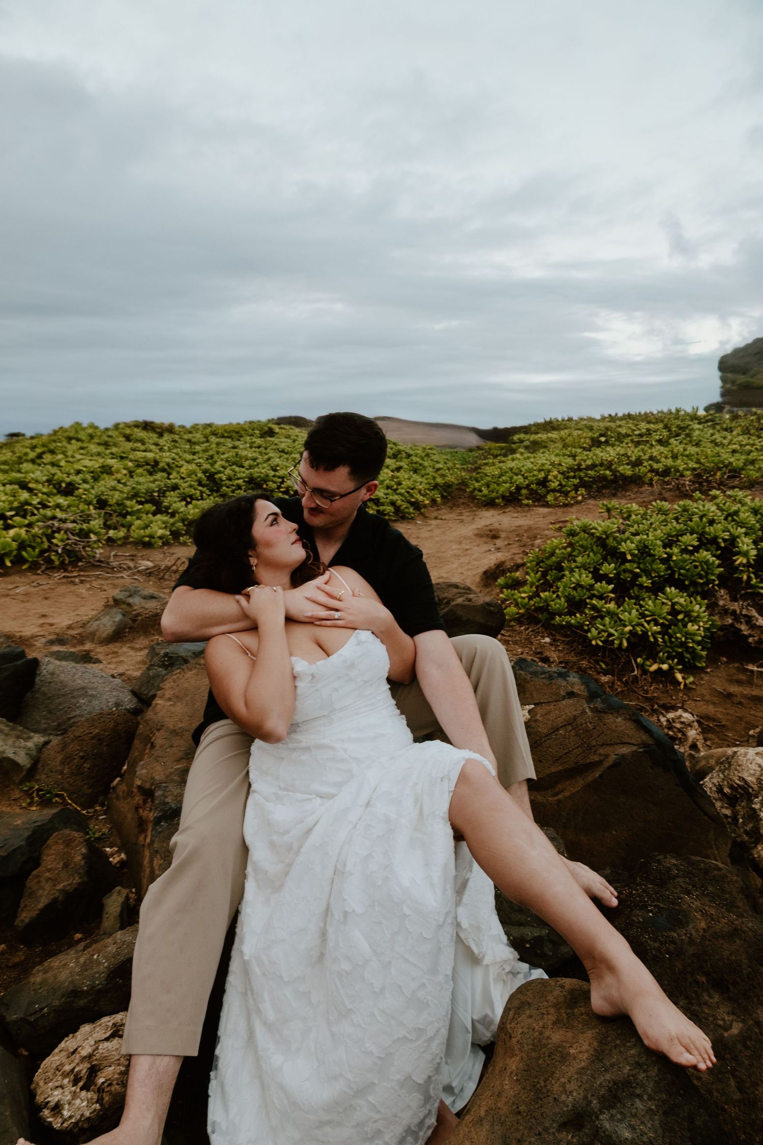 A bride in white lace dress reclines against her groom on coastal rocks with lush greenery behind them.