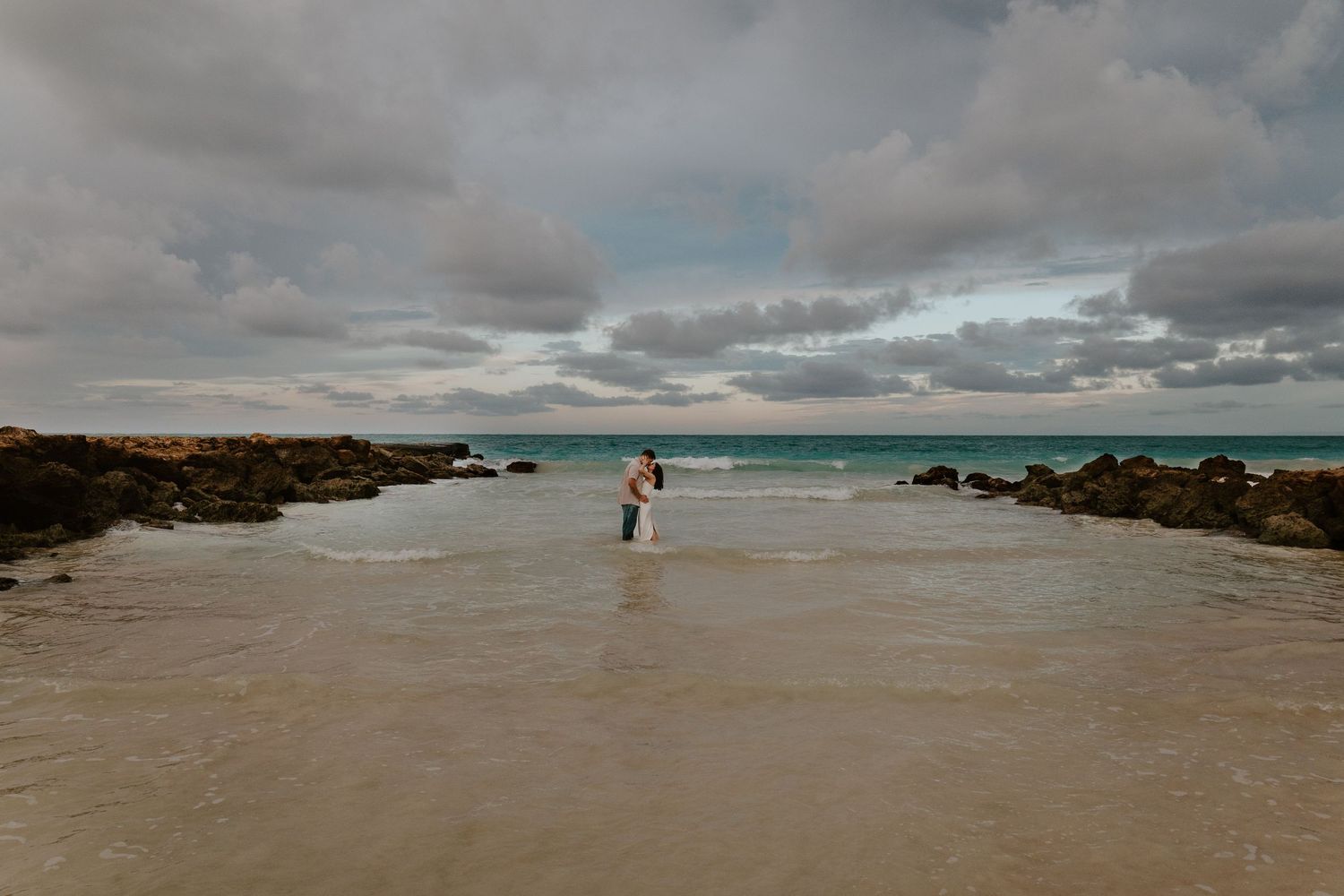 Couple embracing in shallow ocean water on a sandy beach surrounded by rocks under a dramatic cloudy sky.