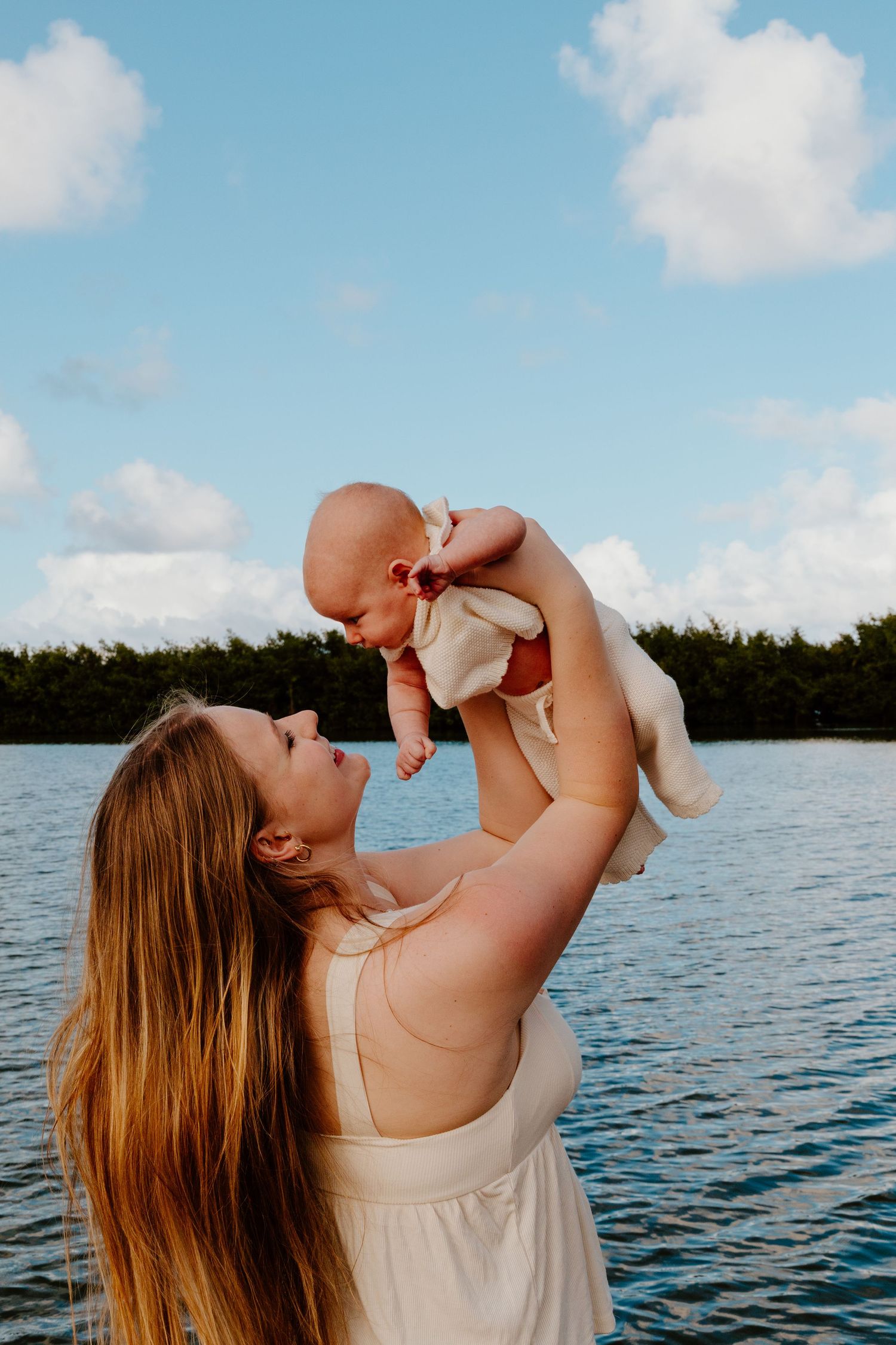 Young mother in white dress lifting smiling newborn baby against blue sky near lake.