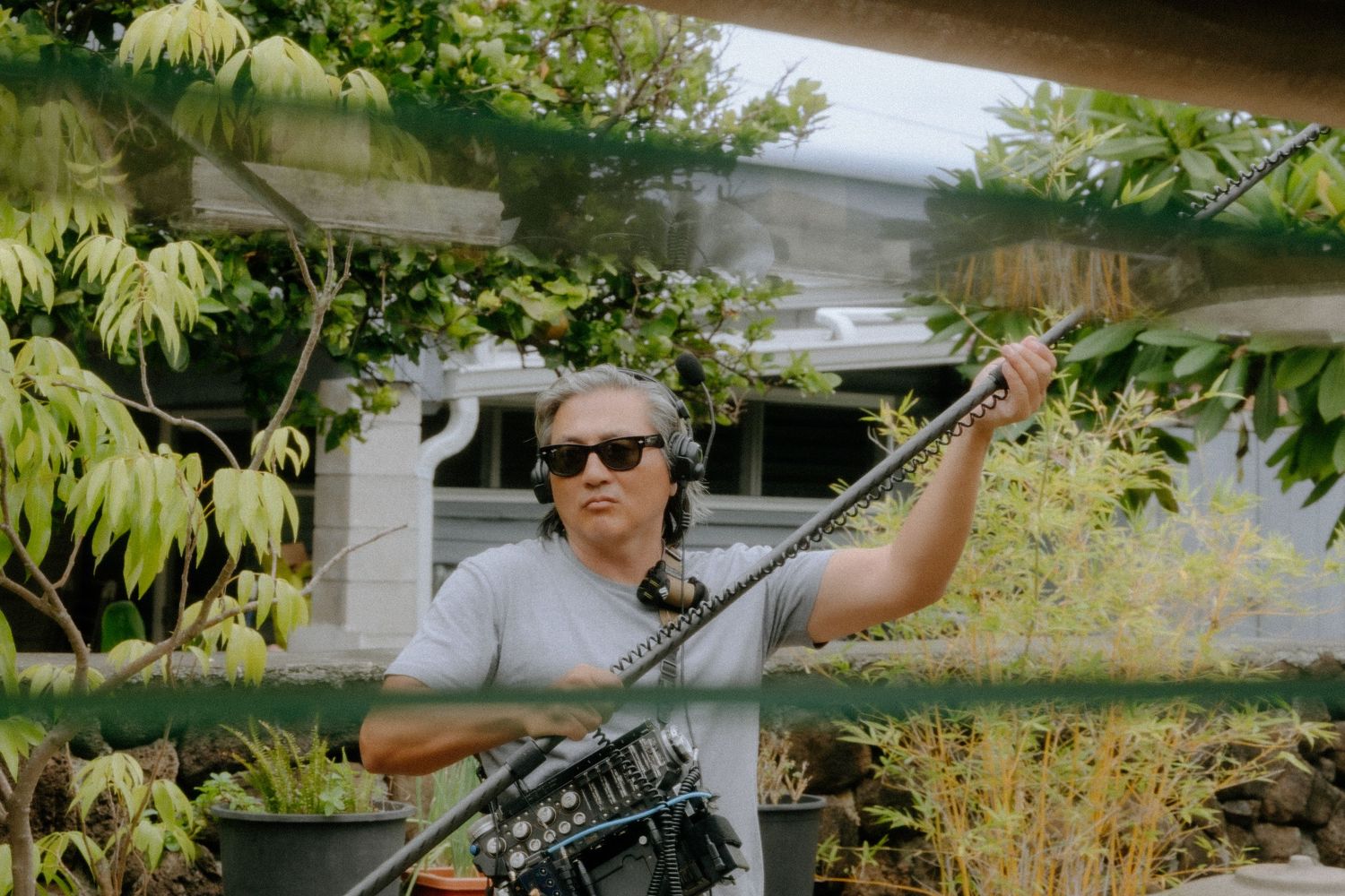 Man in sunglasses and gray shirt trimming hedges with long-handled garden shears outdoors.