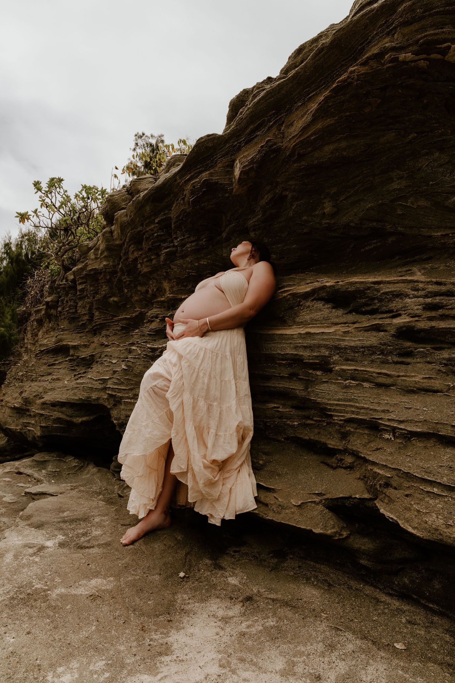 Pregnant woman in a flowing blush dress leaning against rugged rock face, cradling her baby bump outdoors.