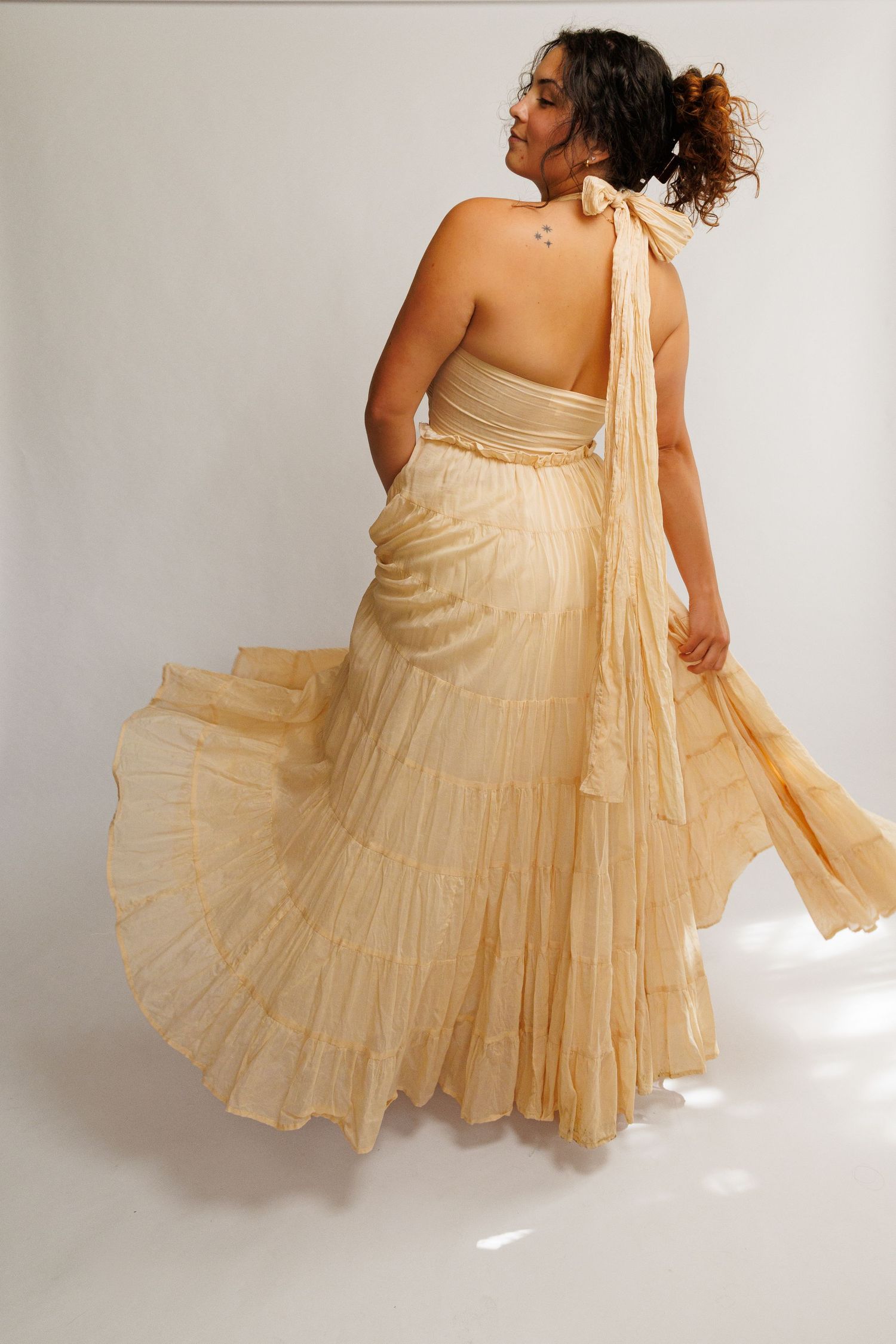 Woman in flowing cream halter gown twirling, showcasing tiered skirt and open back design.