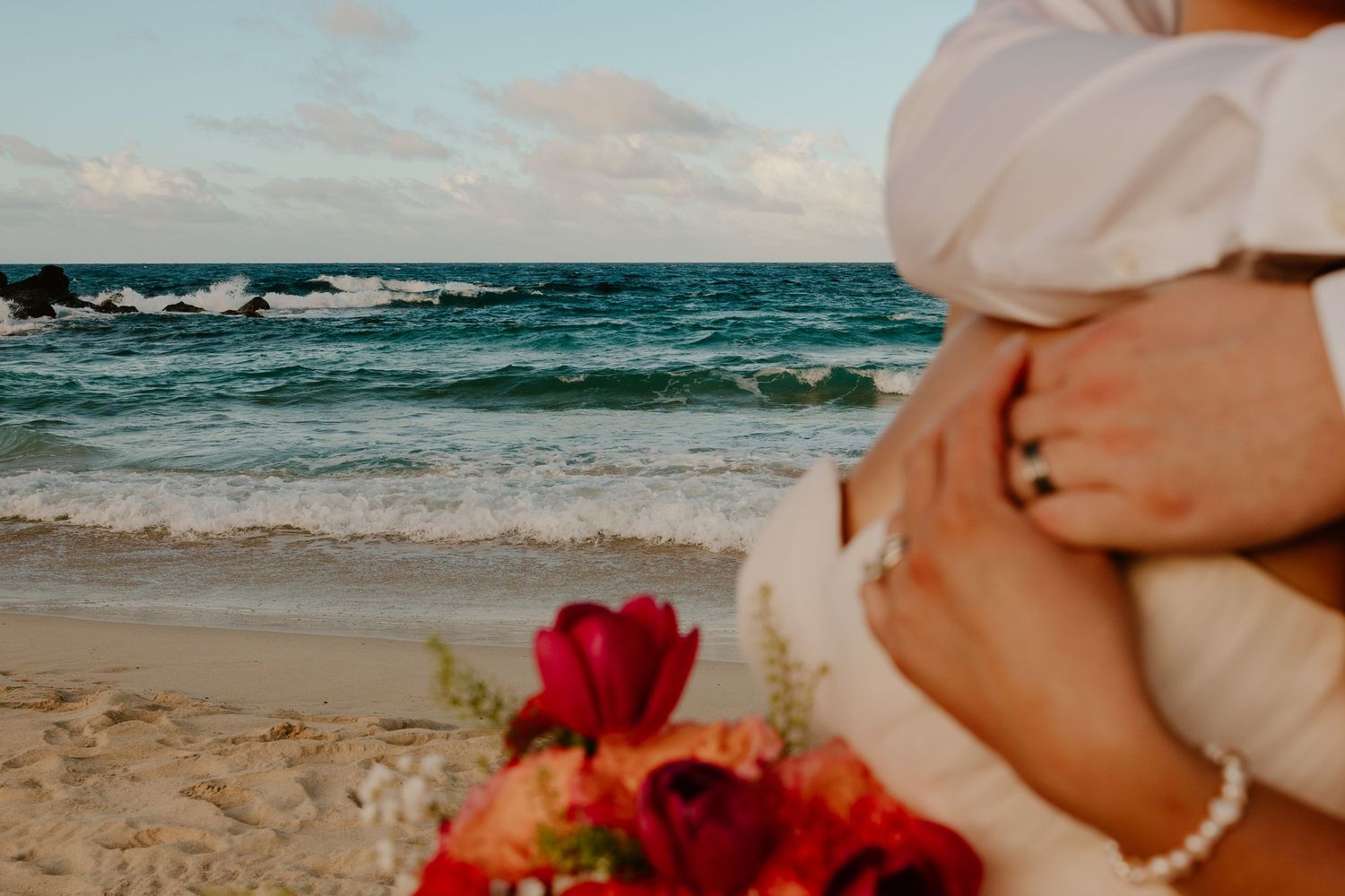 Couple embracing on a sandy beach with turquoise ocean waves and a vibrant red floral bouquet in the foreground.
