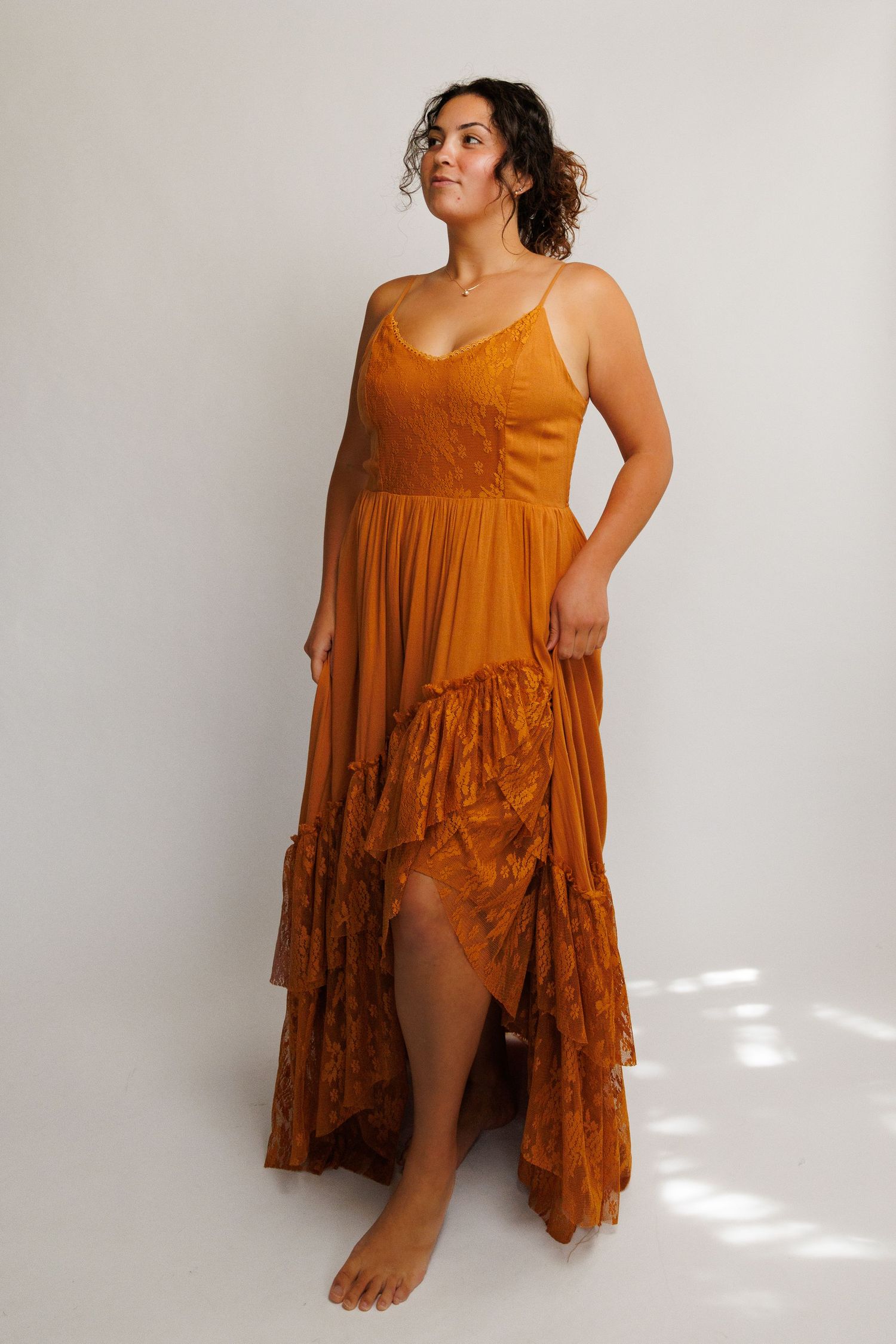 Woman wearing a burnt orange maxi dress with lace details and high-low hem, standing barefoot on white background.