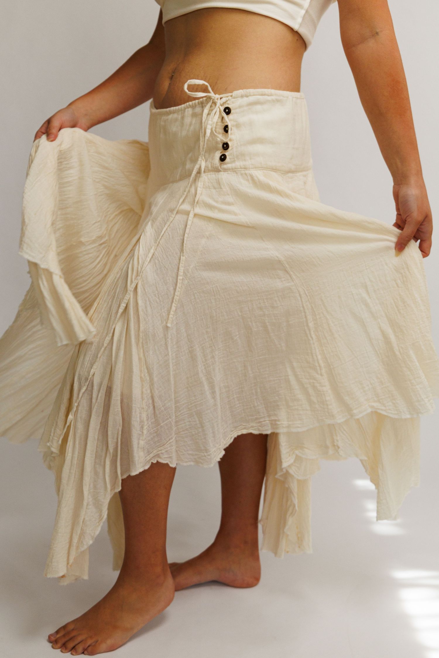 Cream bohemian maxi skirt with lace-up corset waist, flowing layers, and hi-low hem, styled on barefoot model.