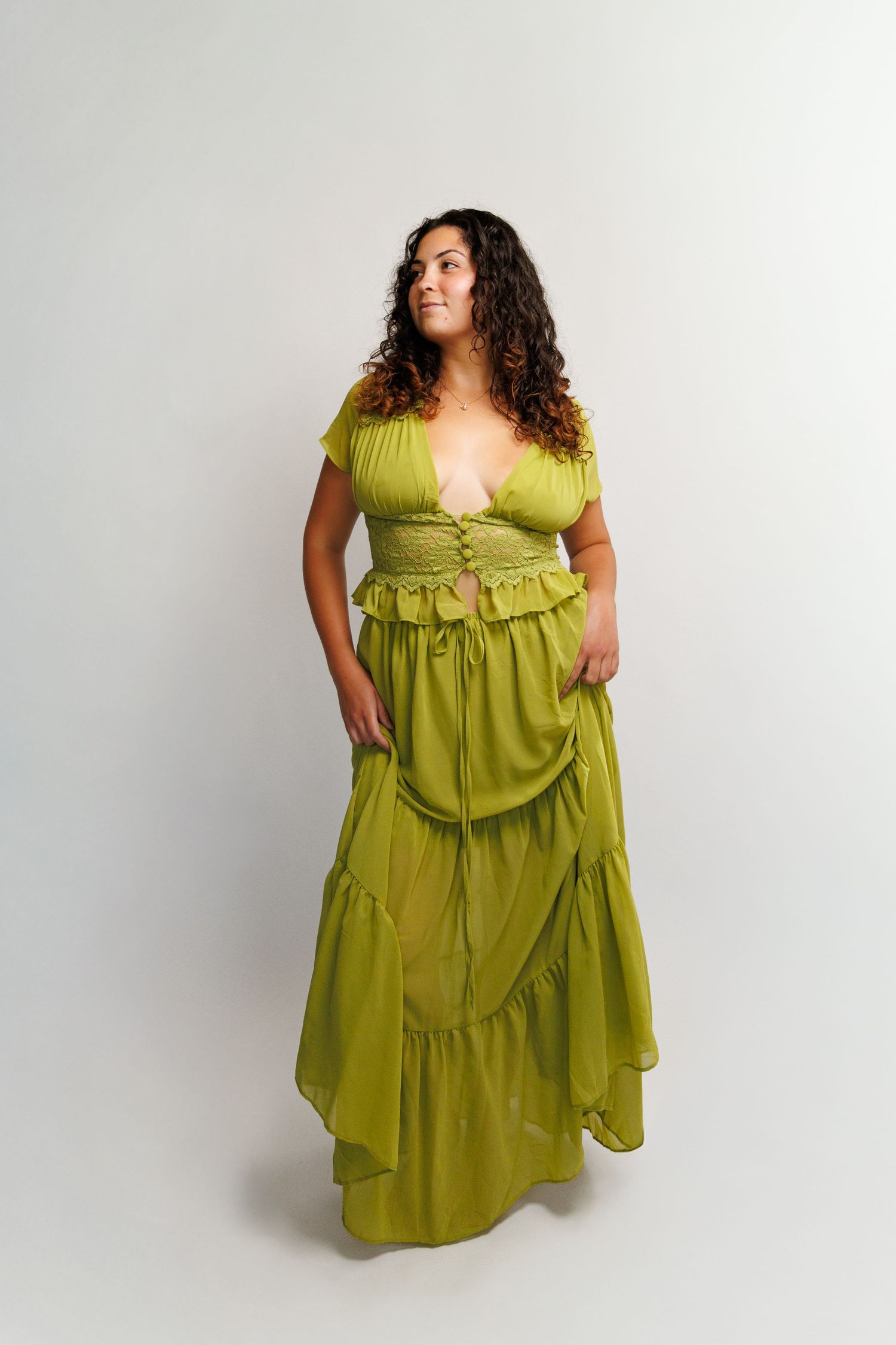 Woman in olive green ruffled maxi dress with deep V-neckline and tiered skirt, posing against white background.