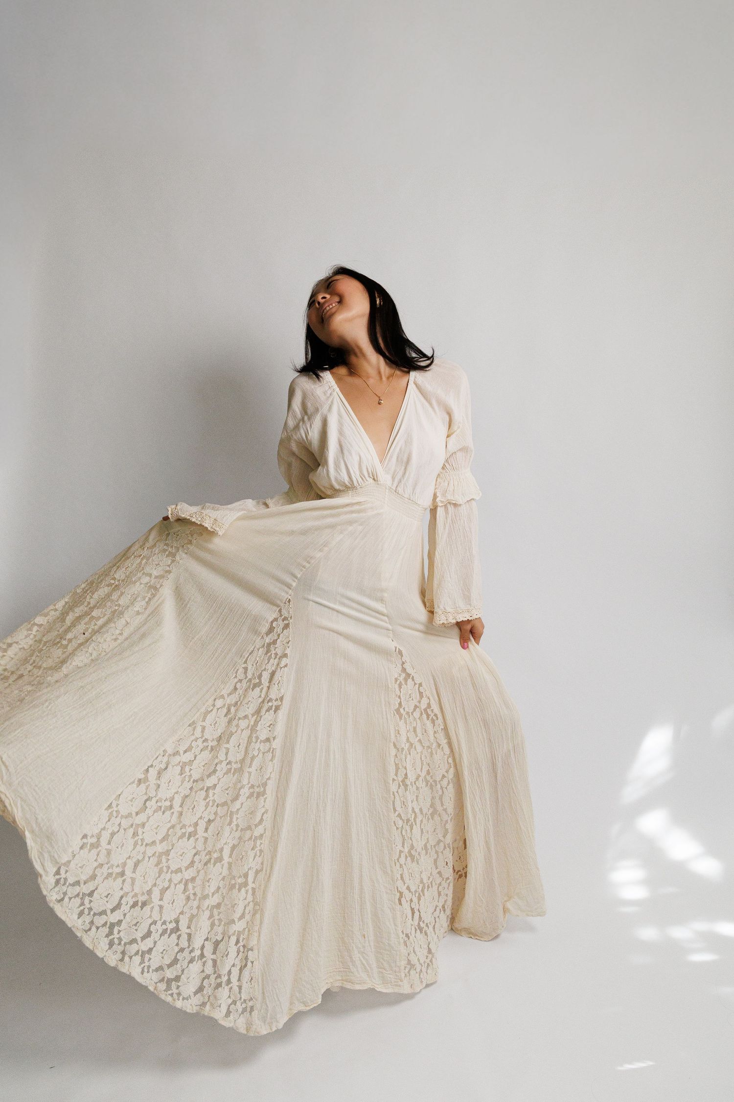 Woman in flowing cream lace boho wedding dress twirling in white studio, long sleeves, V-neck, ethereal bridal gown.