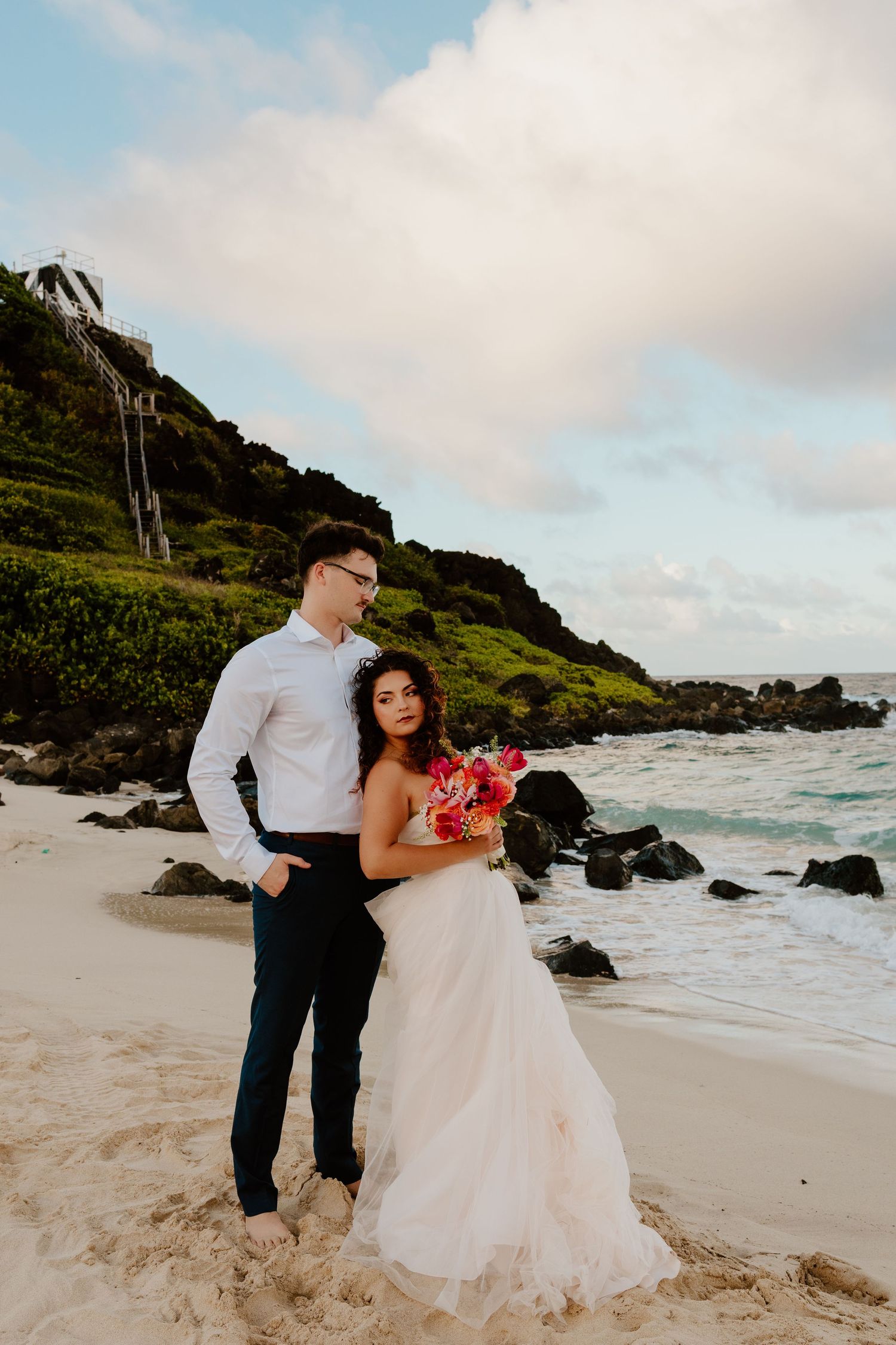 Bride and groom embrace on a sandy Hawaiian beach, she holds a vibrant bouquet, rocky coastline and green hills behind them.