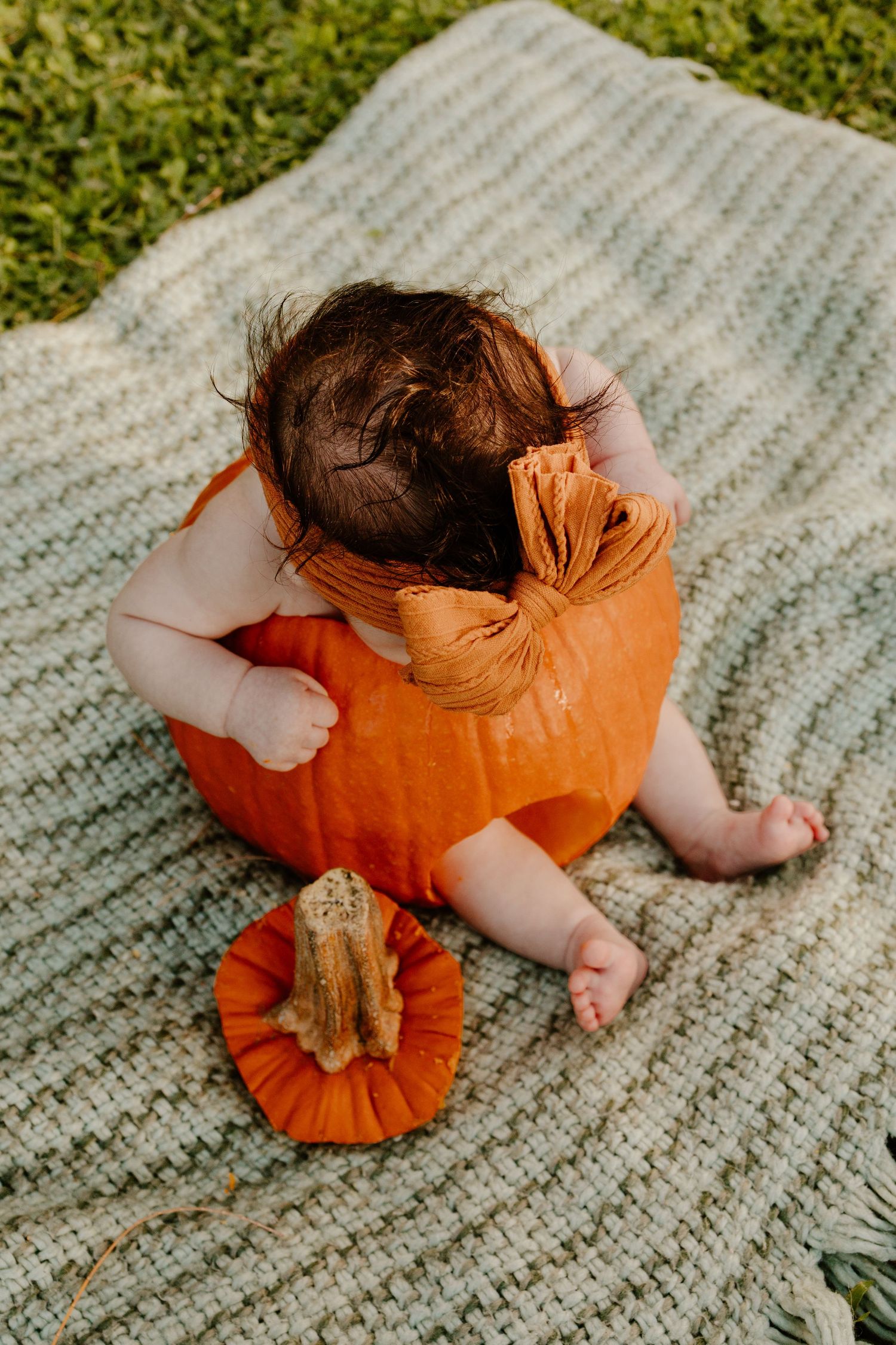 Overhead view of a baby with dark hair sitting inside a hollowed pumpkin on a woven green blanket, with the pumpkin lid beside it.