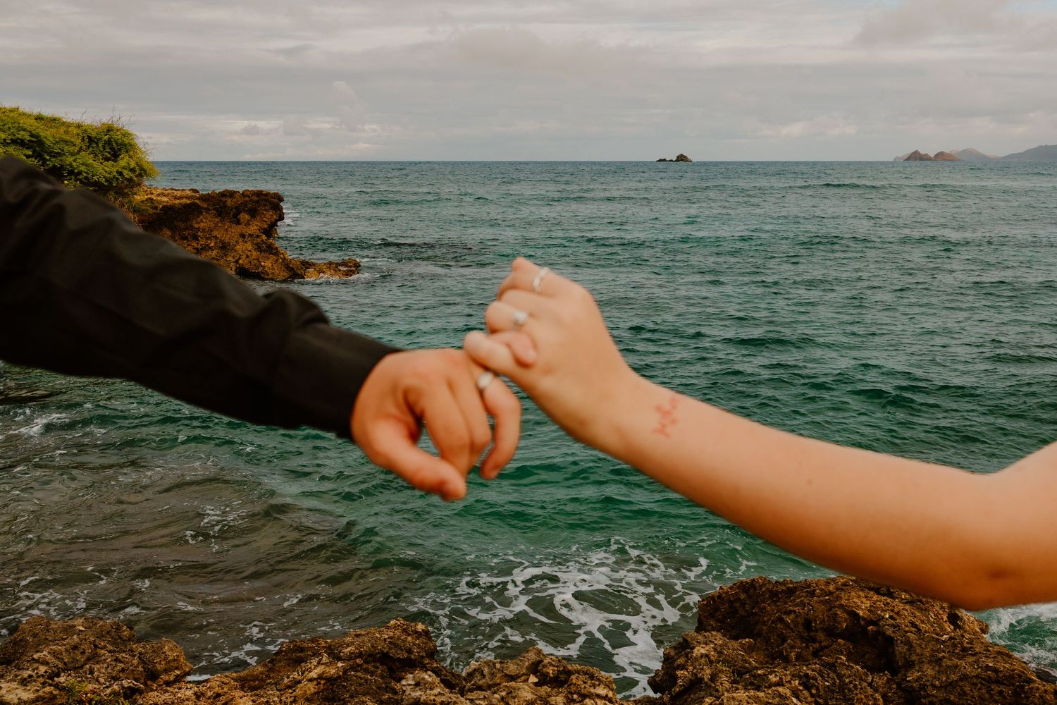 Two people holding hands over rocky coastal shoreline with turquoise ocean waves and small islands in background.