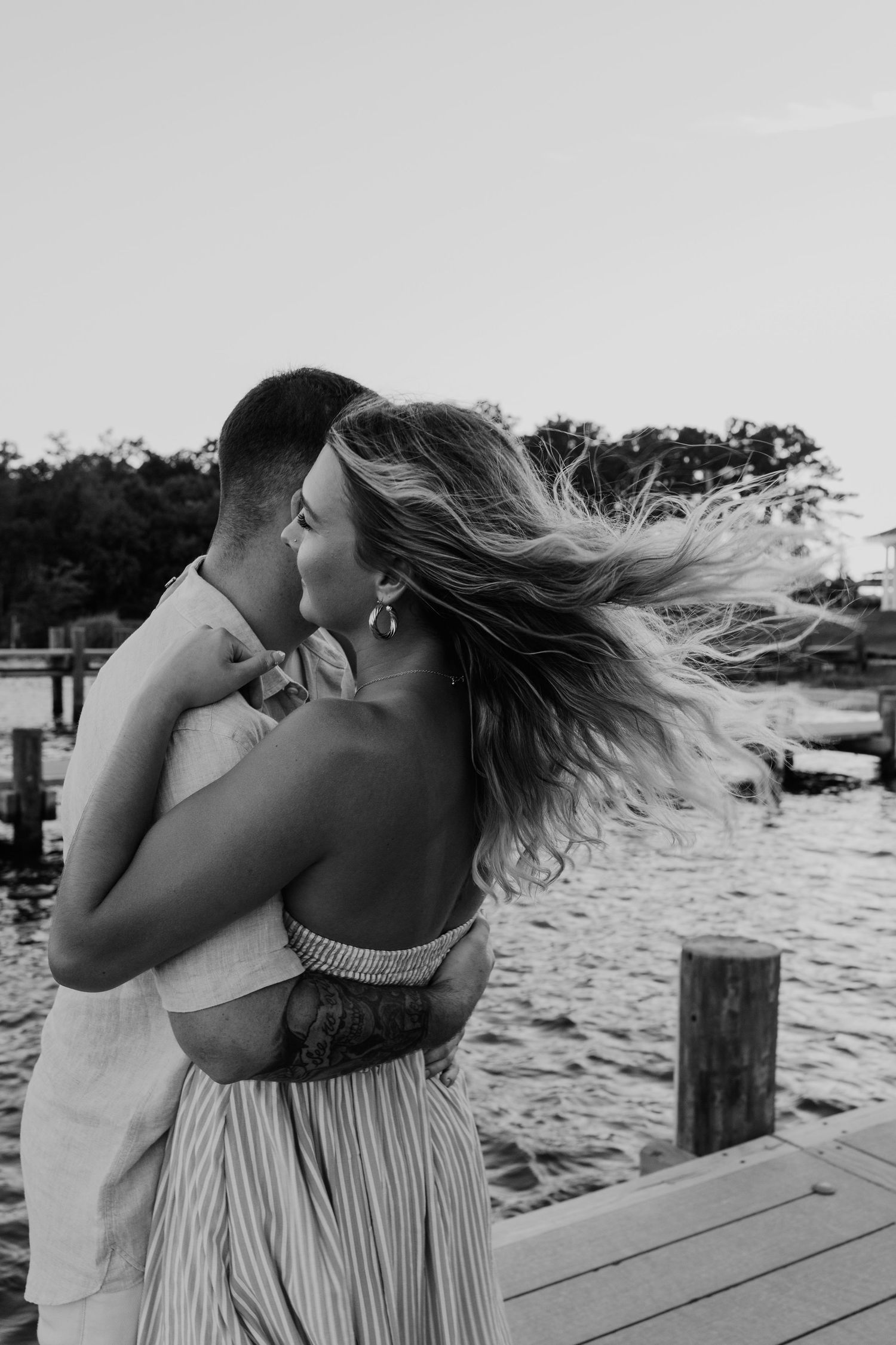 Black and white photo of couple kissing on waterfront dock with womans hair blowing in wind.