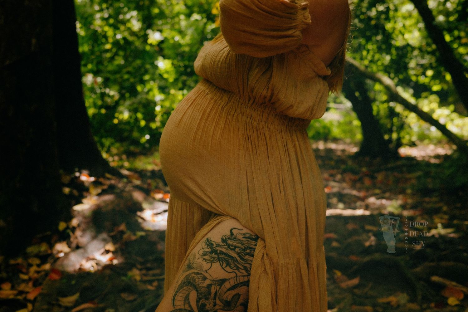 Pregnant woman in golden yellow dress with floral leg tattoo in forest.