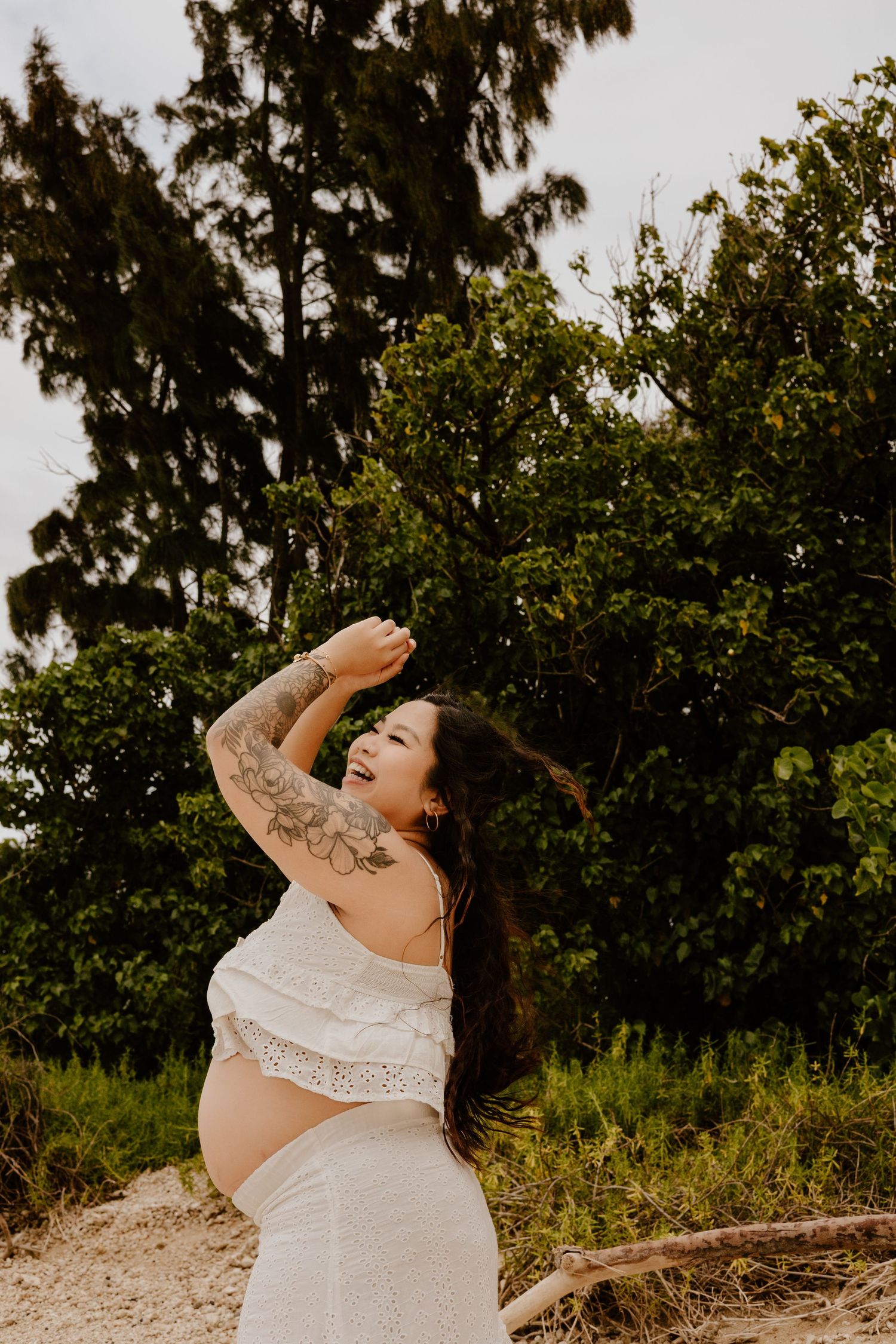 Laughing pregnant woman with sleeve tattoo in white top outdoors near trees.
