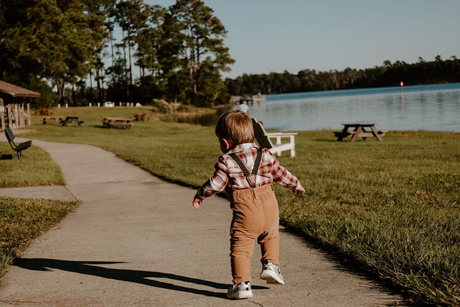 Toddler in plaid shirt and brown overalls walks along a lakeside path surrounded by green grass and tall pine trees.