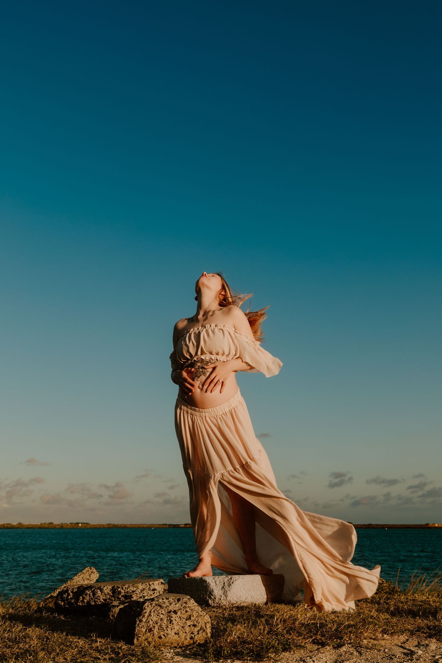 A woman in a flowing peach dress stands on rocks by the ocean, hair blowing in the wind under a clear blue sky.