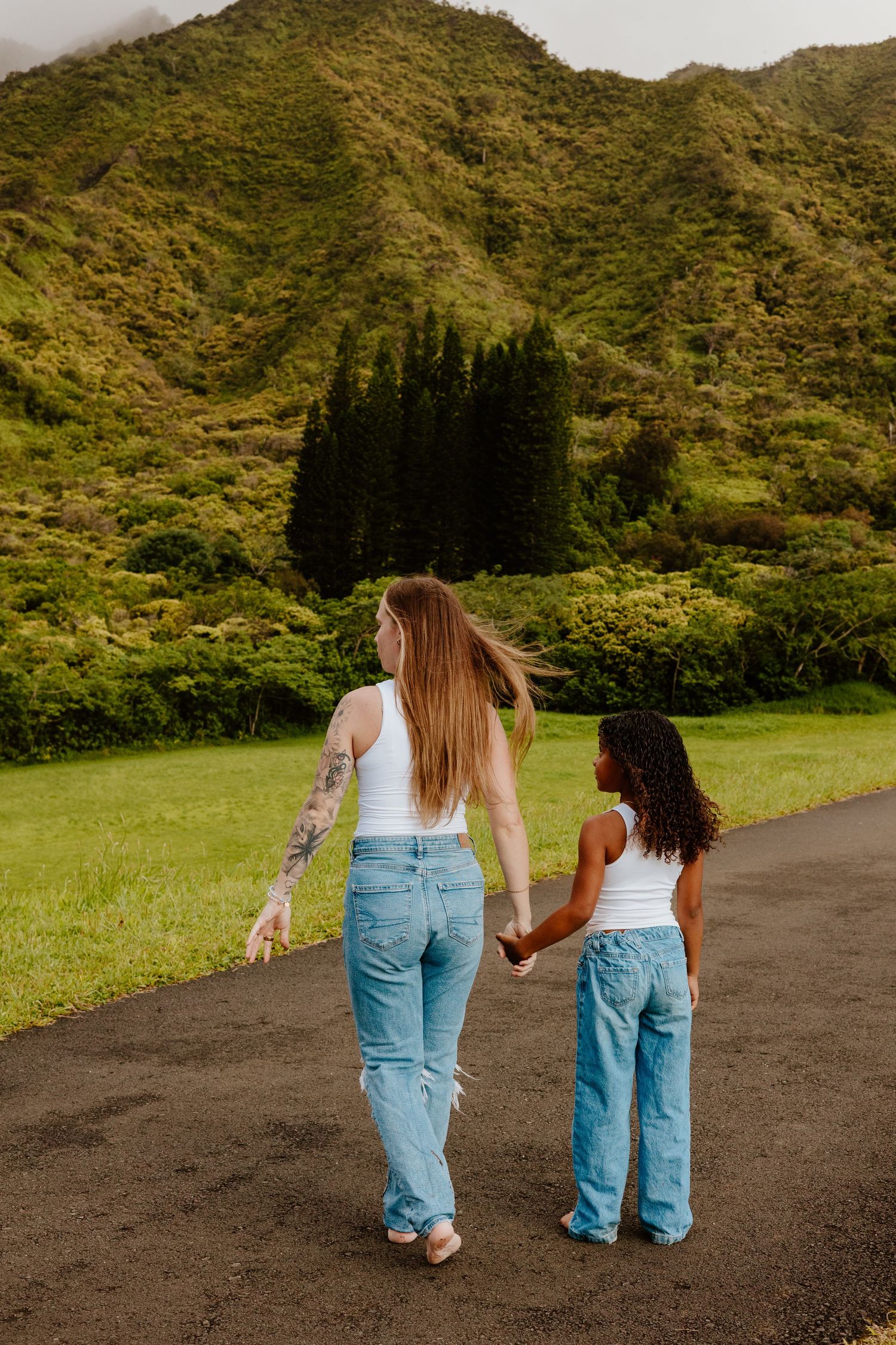 Two females in white tops and jeans walking hand in hand toward lush green mountains.