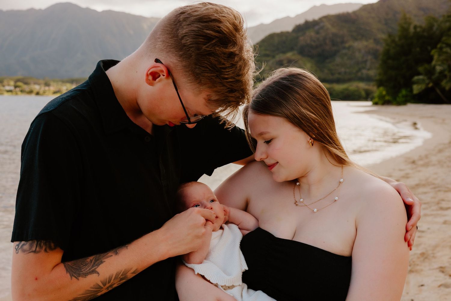 Young couple with tattoos tenderly holding their newborn baby on a sandy beach.