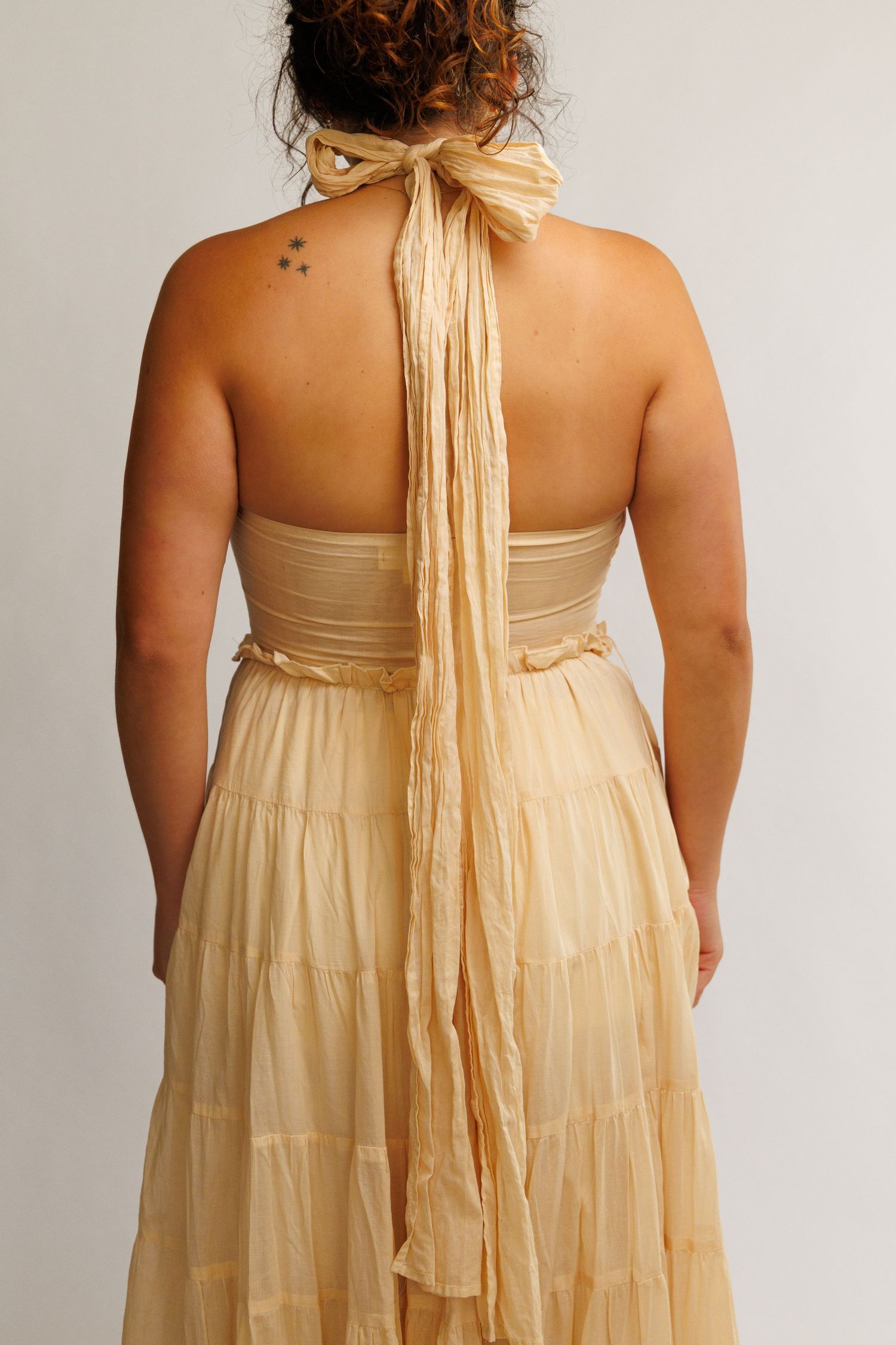 Woman wearing a backless golden halter maxi dress with tiered skirt and long bow tie detail.