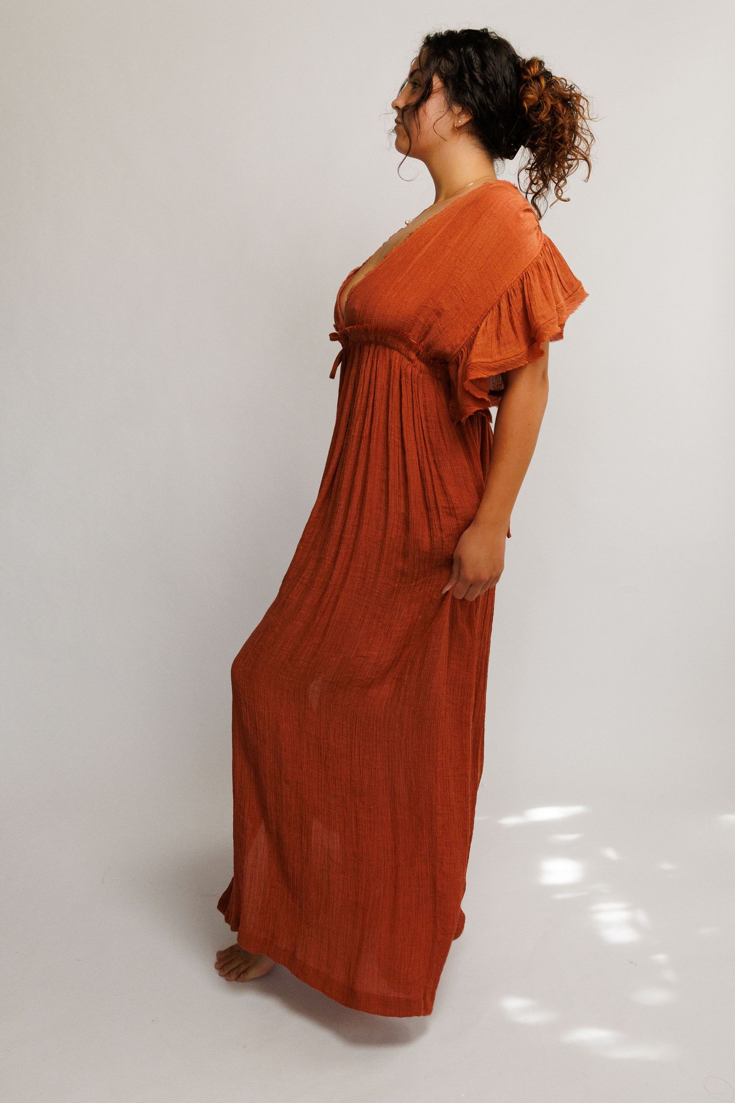 Woman wearing a rust orange maxi dress with flutter sleeves and empire waist, posing sideways in a studio.
