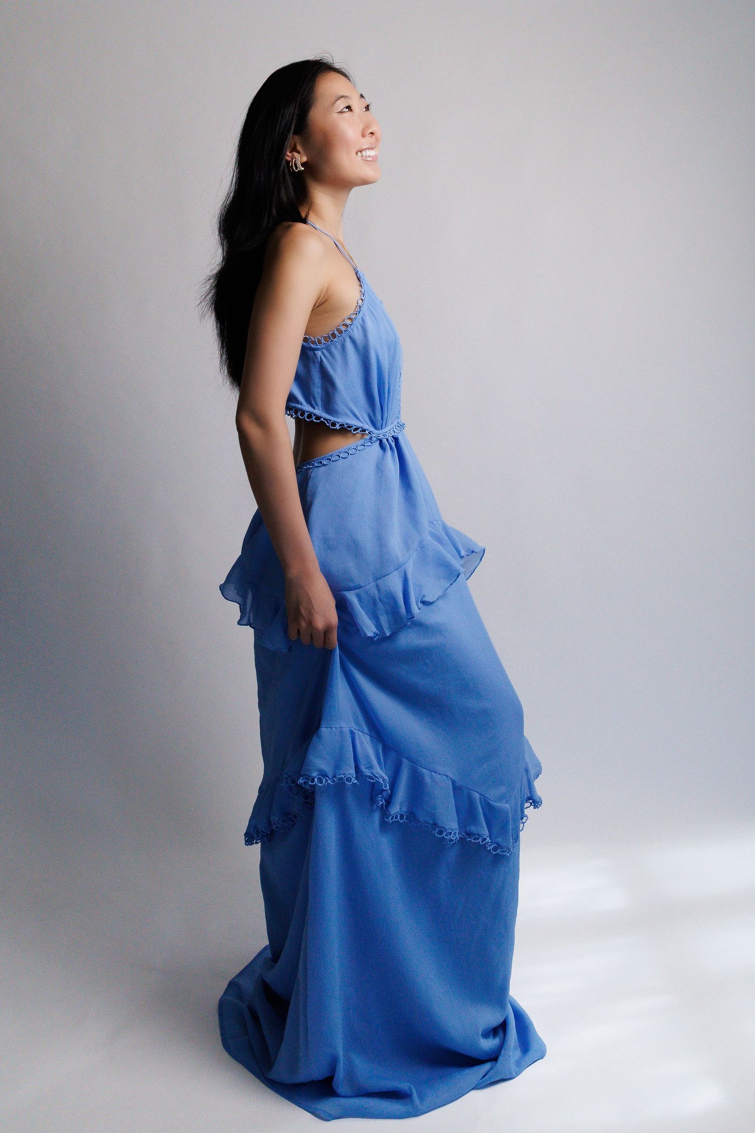 Woman in blue ruffled maxi dress with cutout waist, smiling sideways in studio.