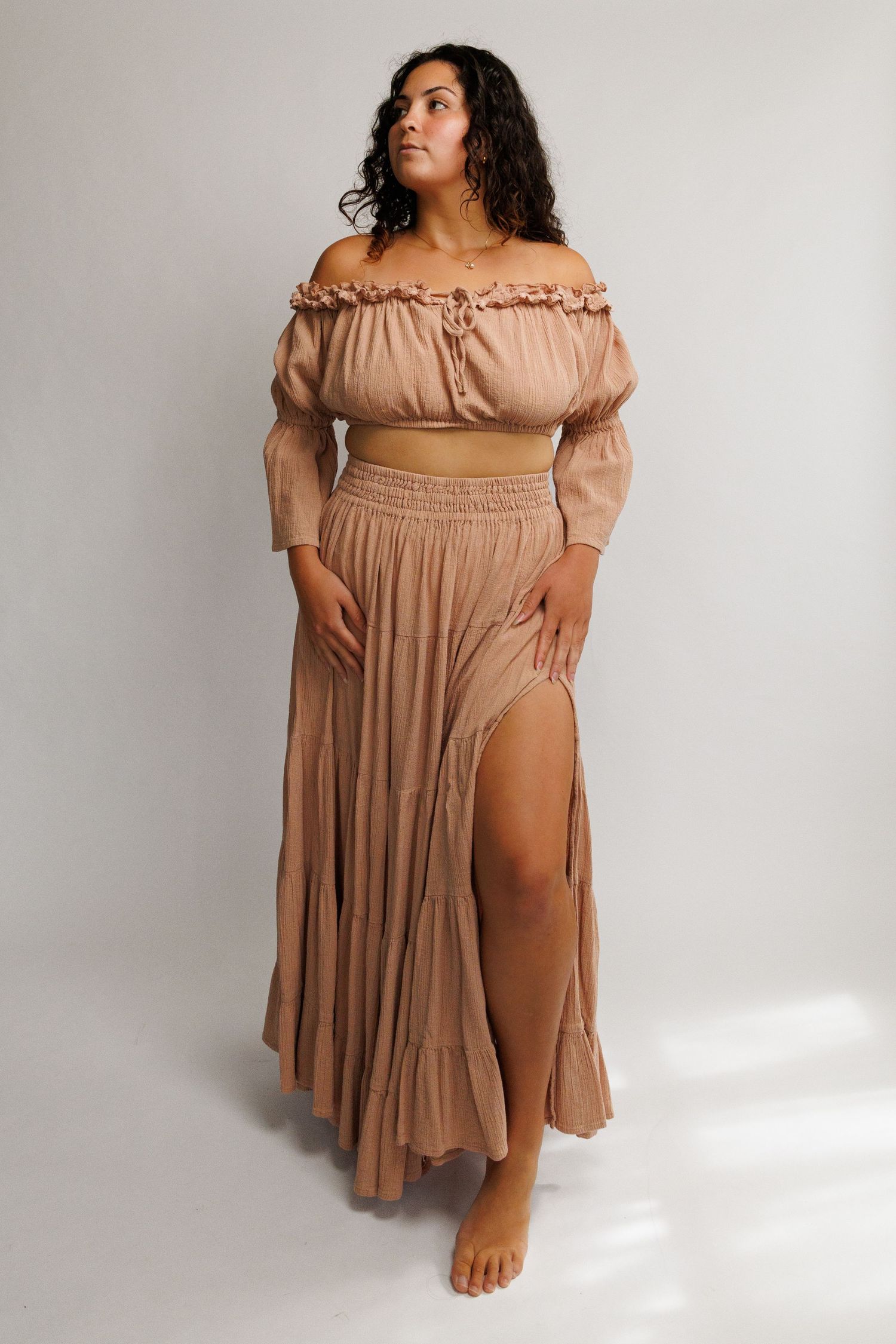 Woman wearing a tan off-shoulder crop top and matching maxi skirt with a front slit, posing against a white background.