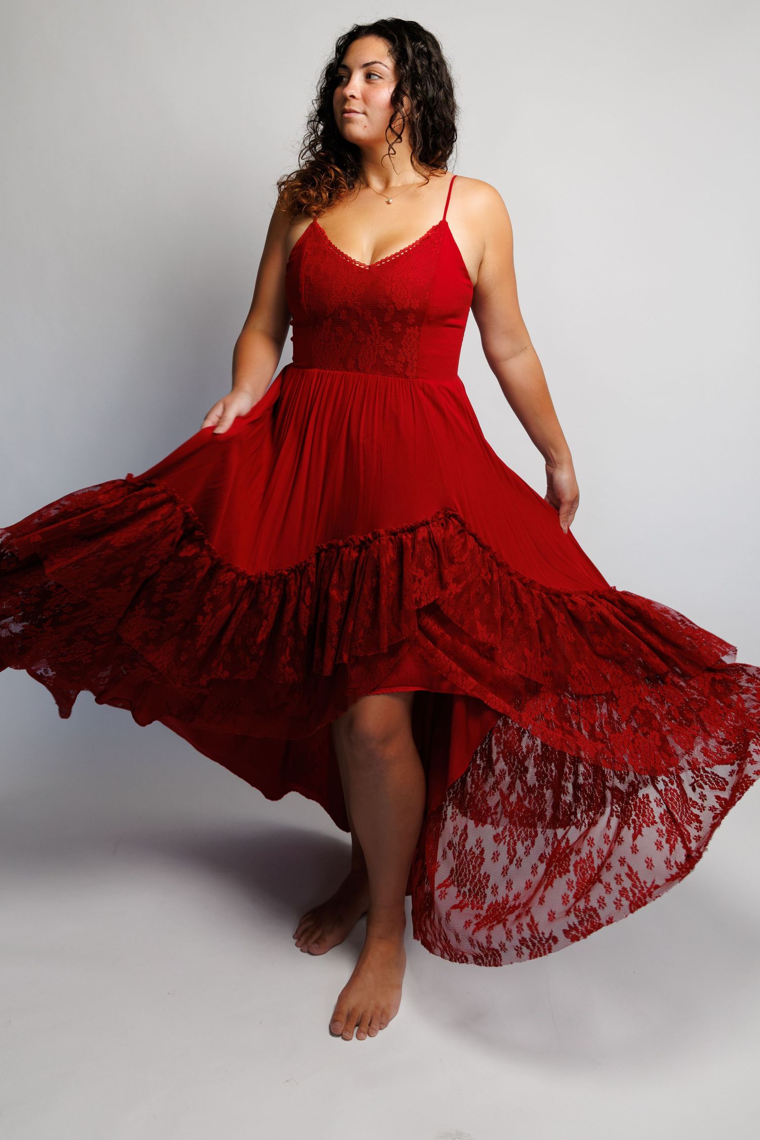 Woman in flowing red lace high-low dress with spaghetti straps, twirling barefoot against a gray background.