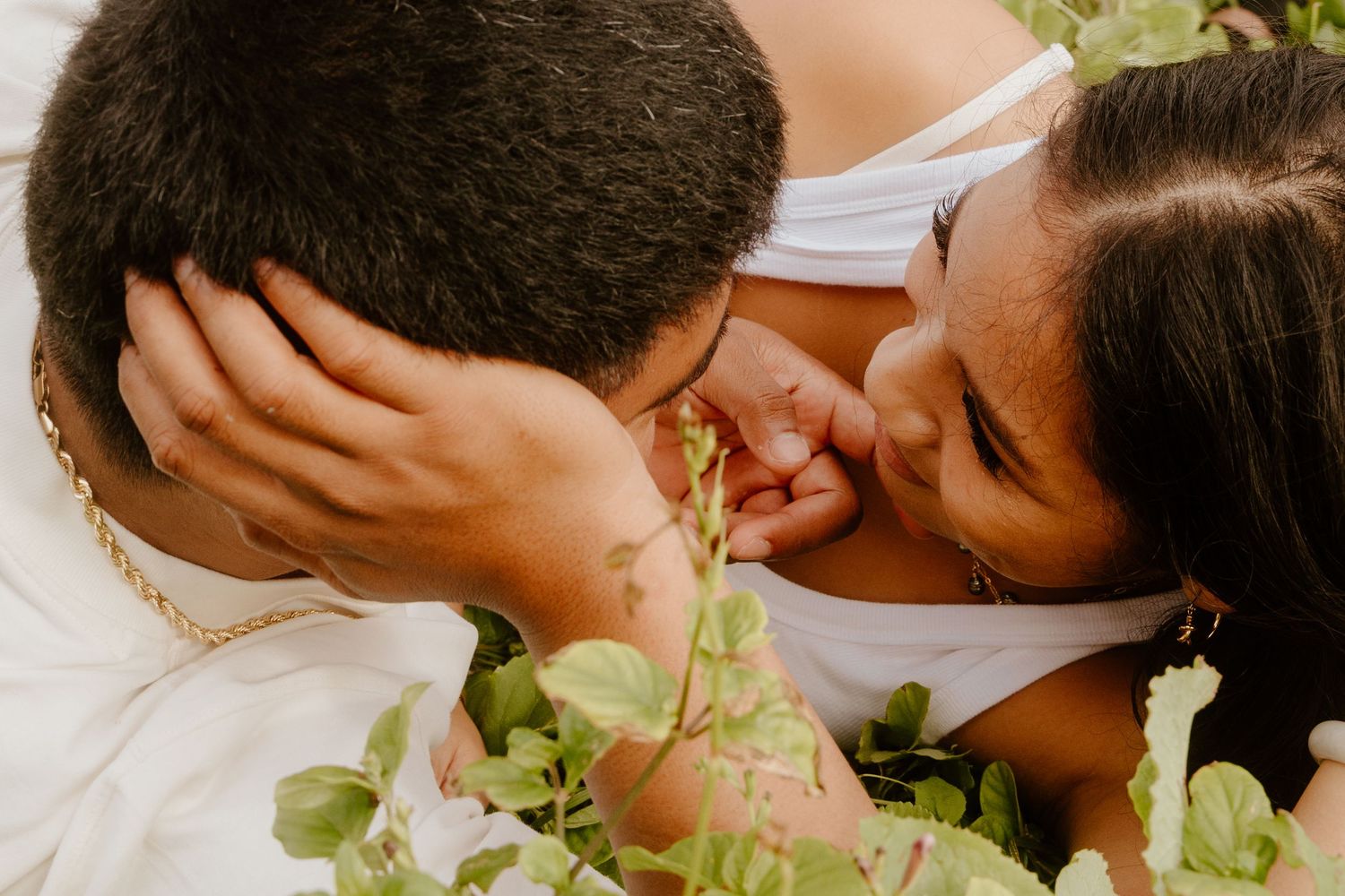 A close-up of a couple laughing and kissing while lying among lush green tropical plants.