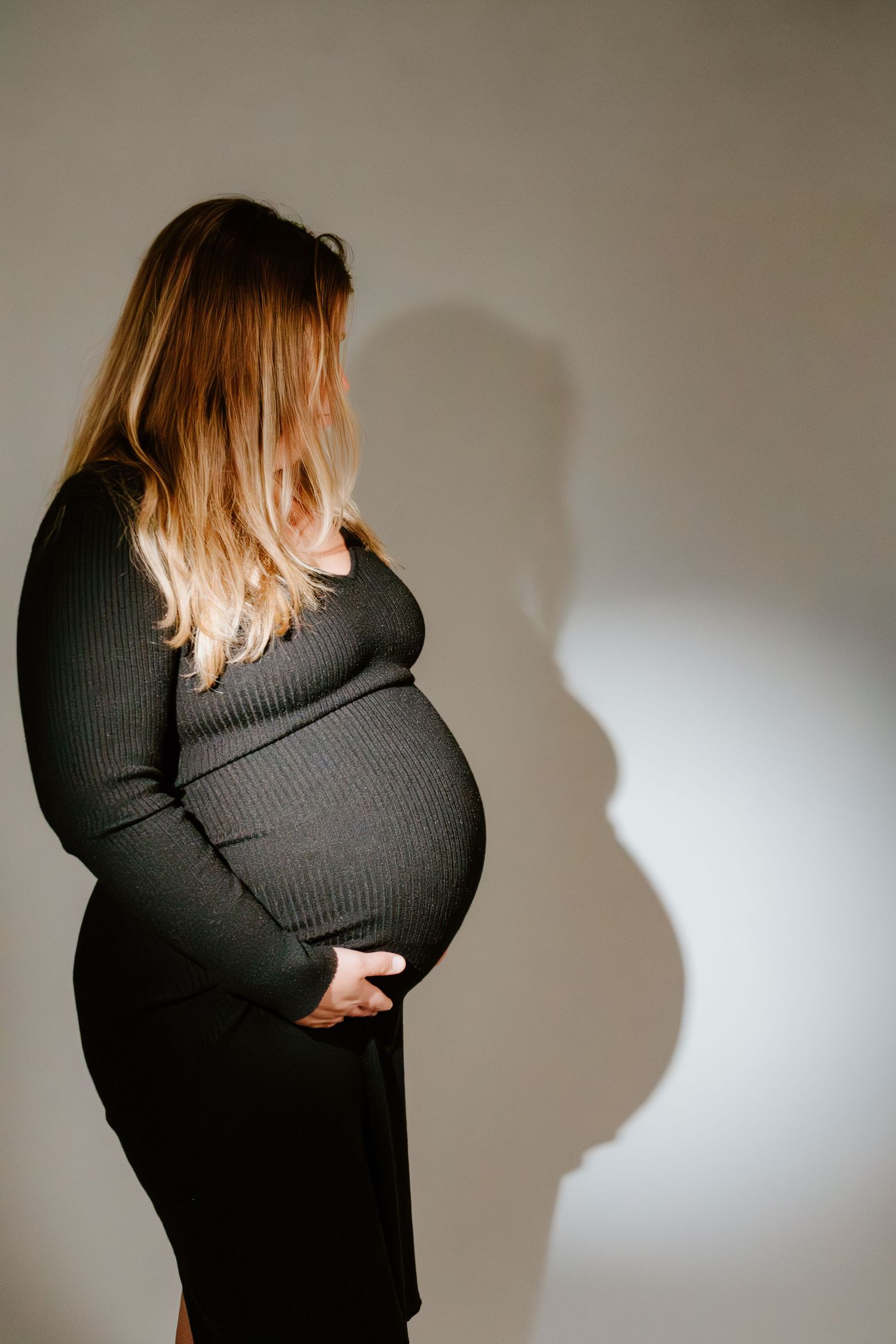 Pregnant woman in dark ribbed dress holding baby bump sideways, casting dramatic shadow on wall.