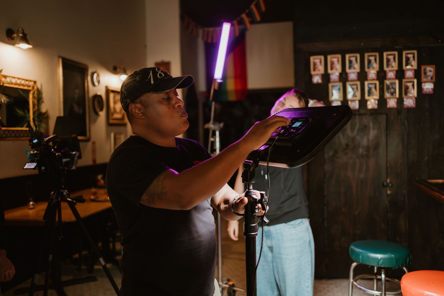 A tattoo artist in a black cap applies a stencil to a client in a dimly lit tattoo parlor with colorful neon lights.