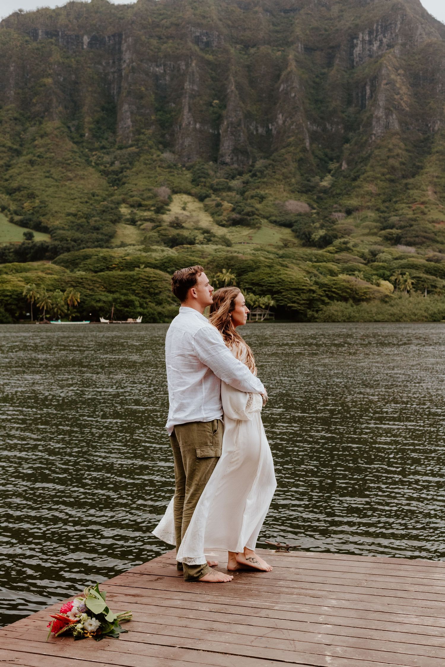 Couple embracing on a wooden dock with lush green mountains and calm water in Hawaii.