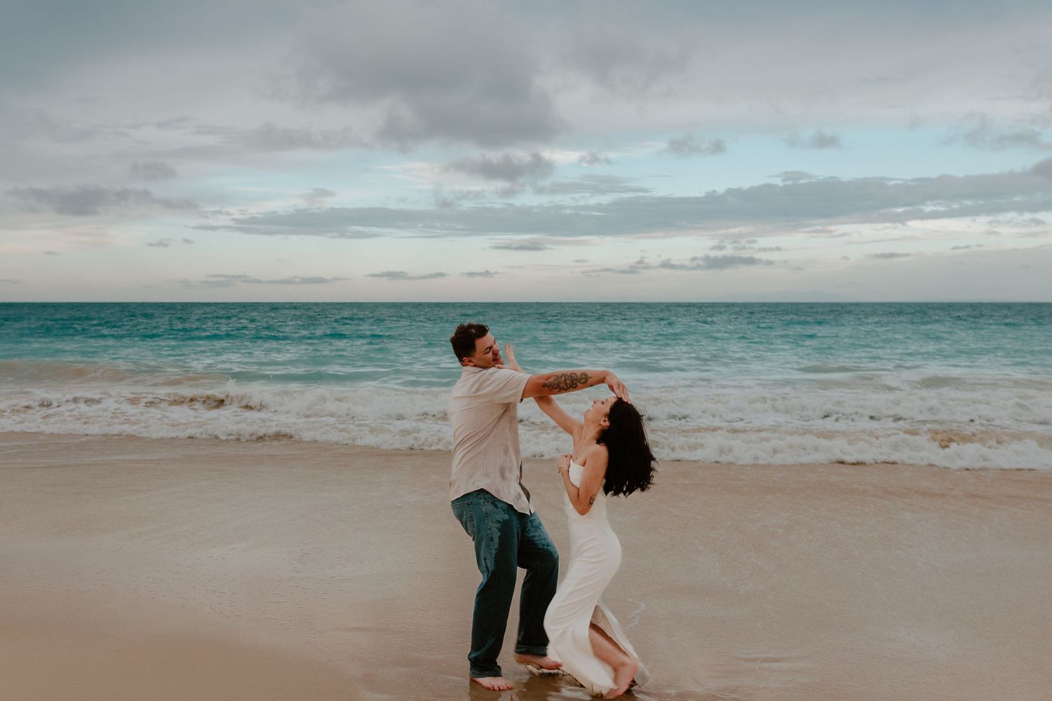 Couple dancing and spinning on a tropical beach at dusk with turquoise ocean and cloudy sky behind them.