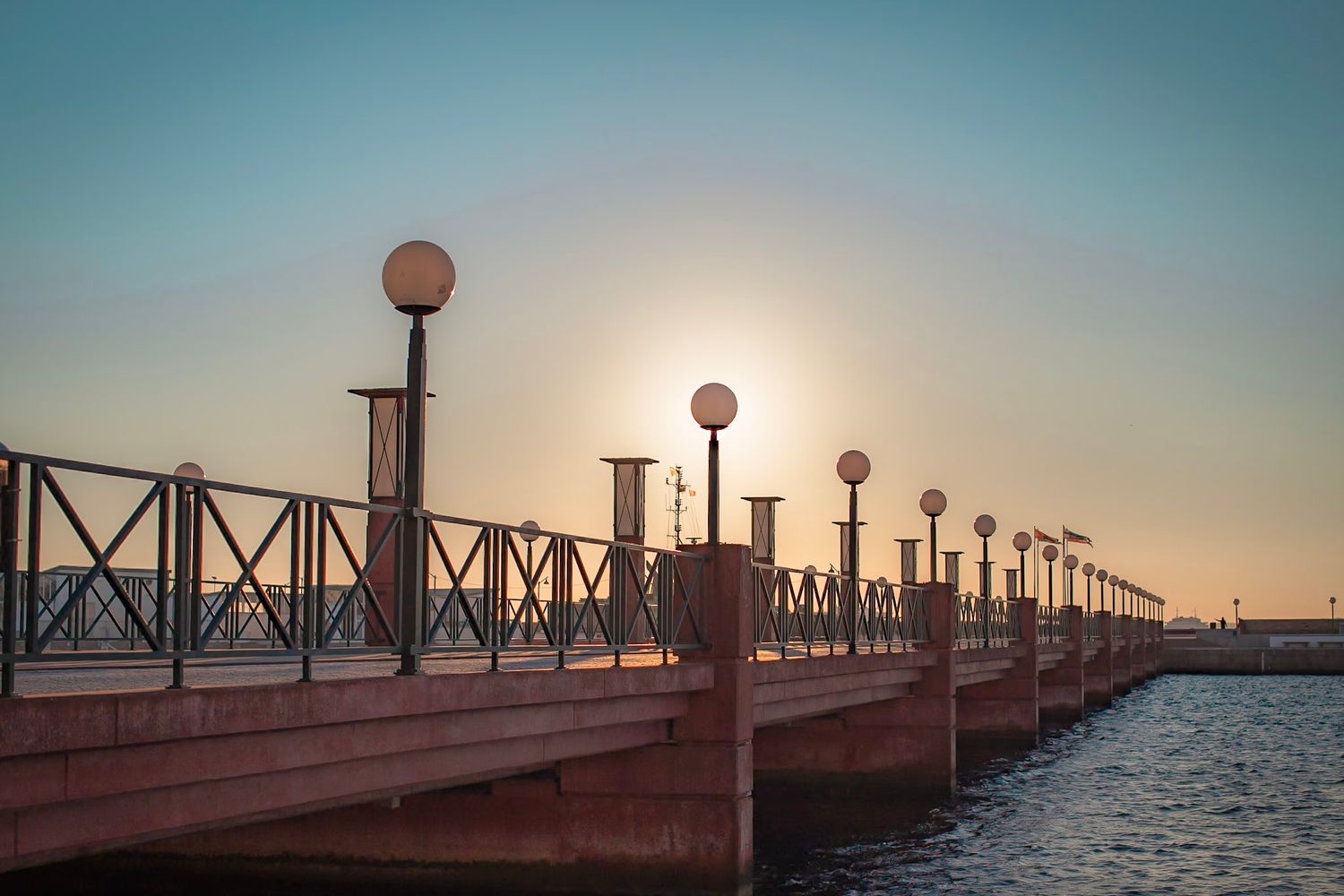 A scenic pier with ornate lampposts extends into the water during a tranquil sunset creating a peaceful coastal view.