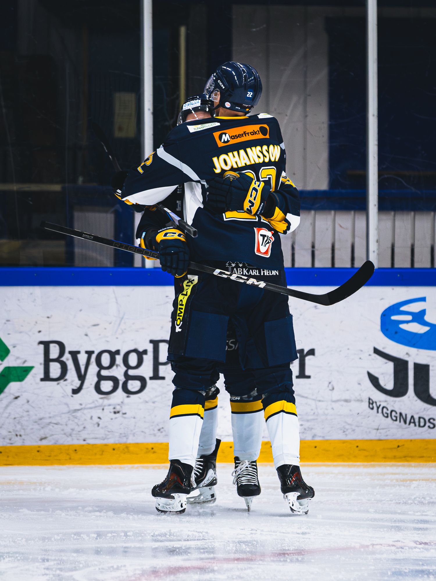 Players in black uniforms skate on ice rink with advertising boards visible in background.