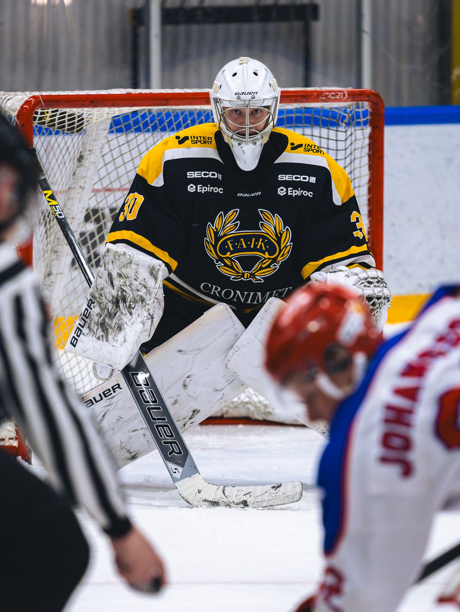 Goalie in black uniform defends the net against opposing team in red and white.
