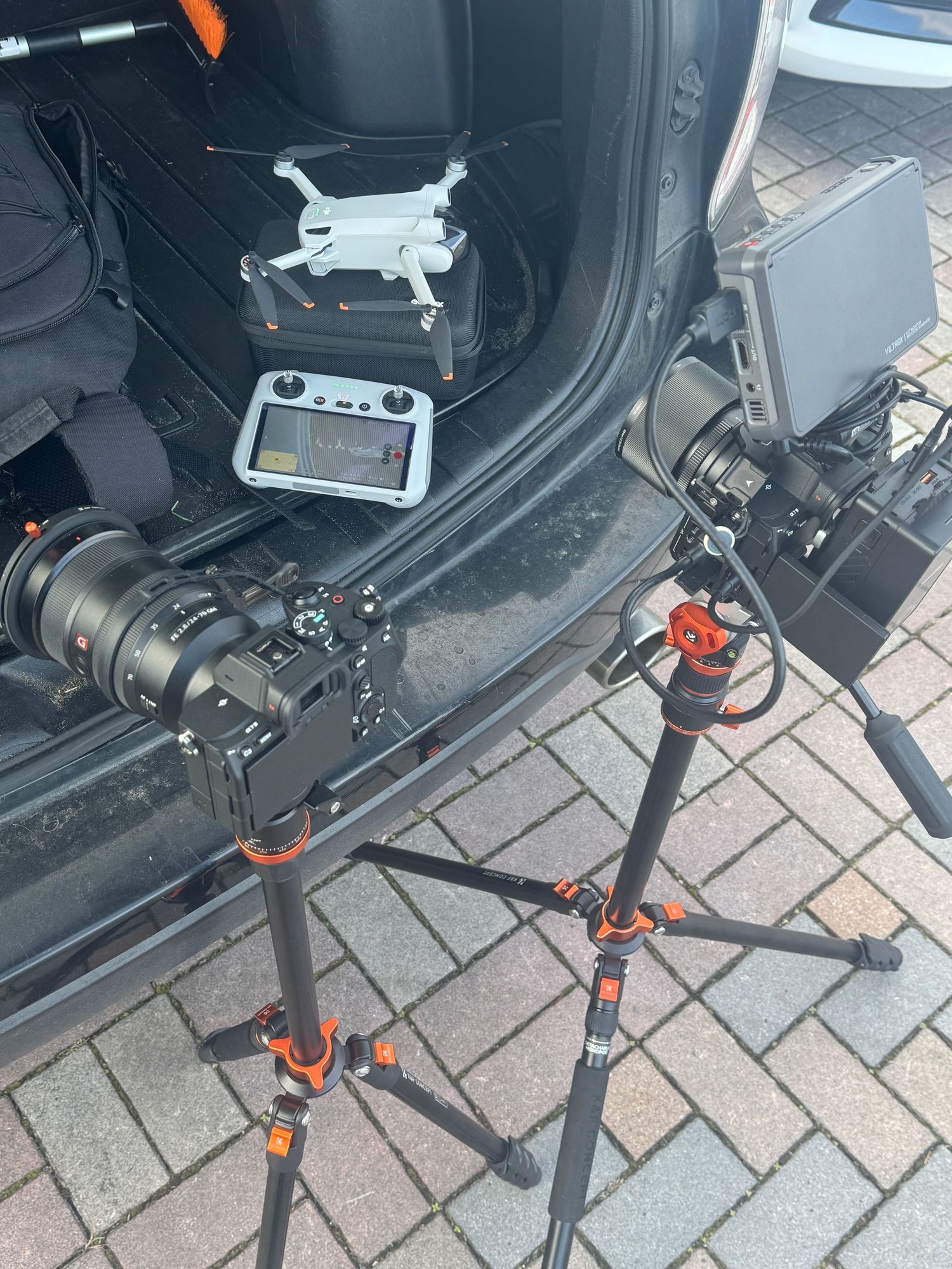 Professional camera equipment on tripods with a DJI drone and controller in a car trunk, ready for a shoot.