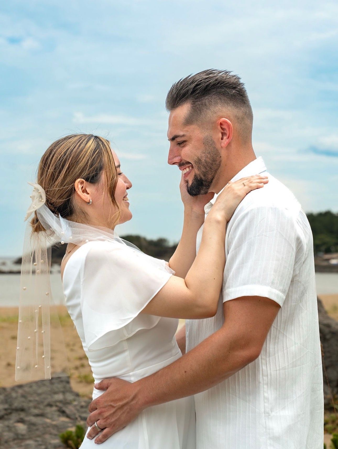A couple in white outfits embraces lovingly on a rocky beach during a romantic outdoor photoshoot.