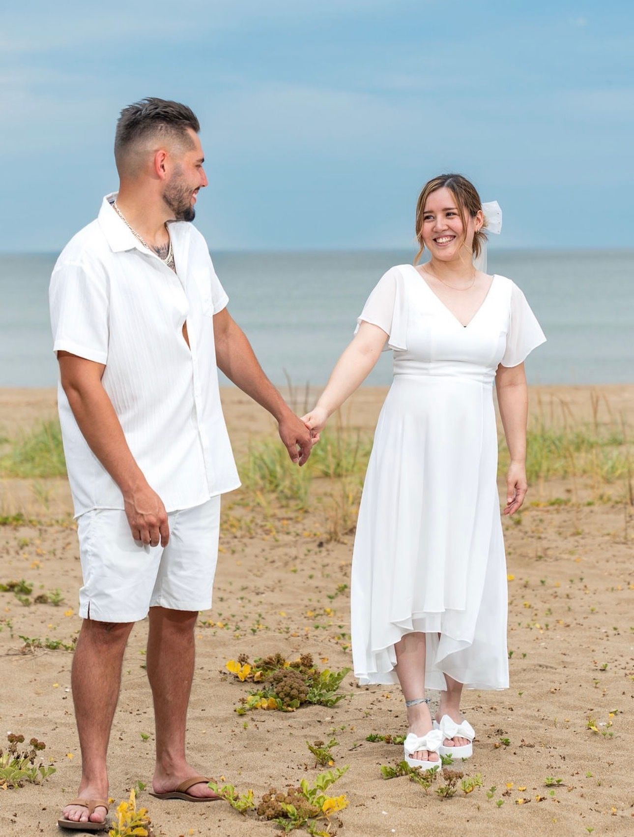 Couple in white summer outfits walking barefoot on sandy beach.