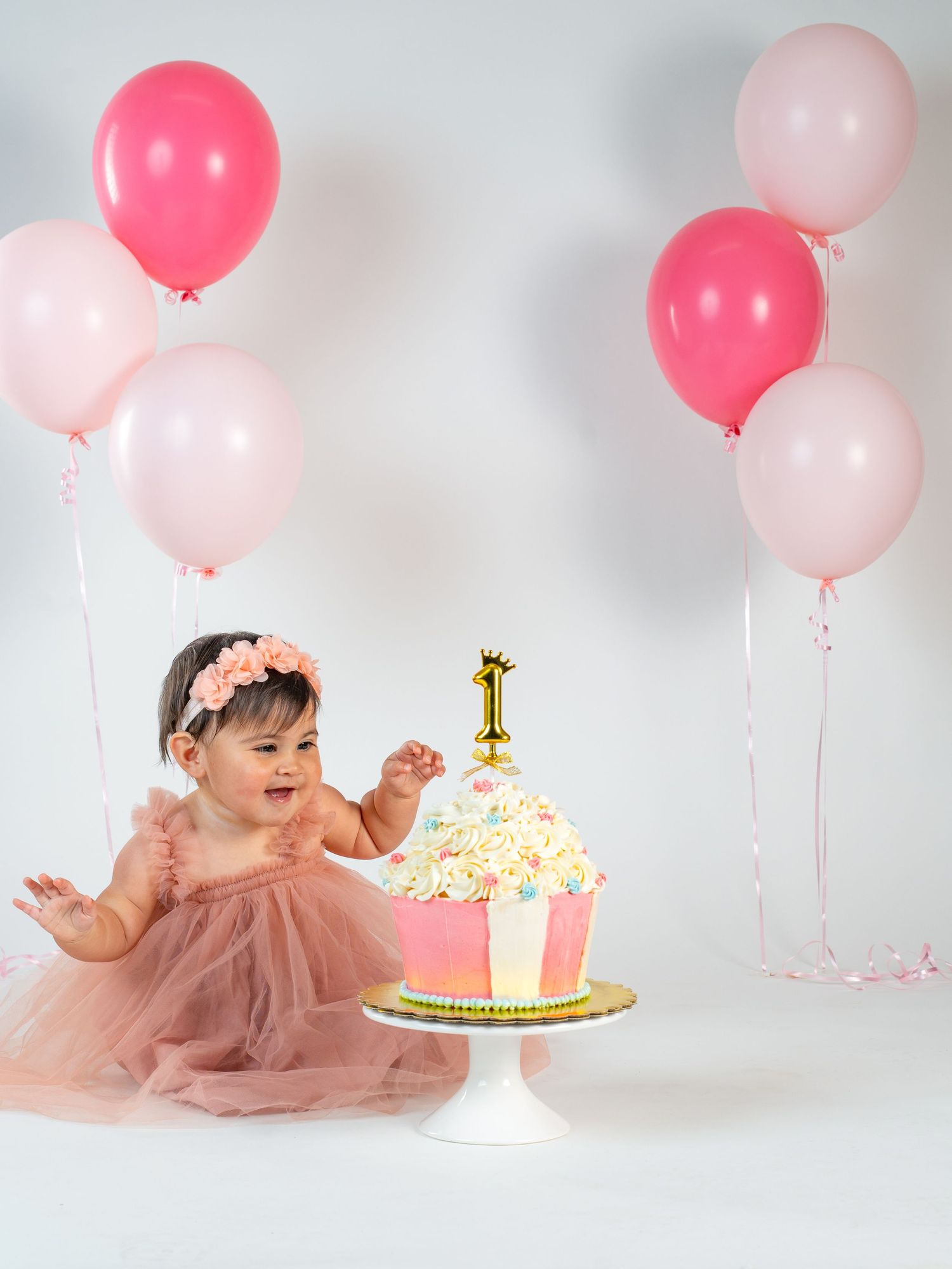A baby in a pink tulle dress sits beside a first birthday cake with pink and coral balloons in the background.