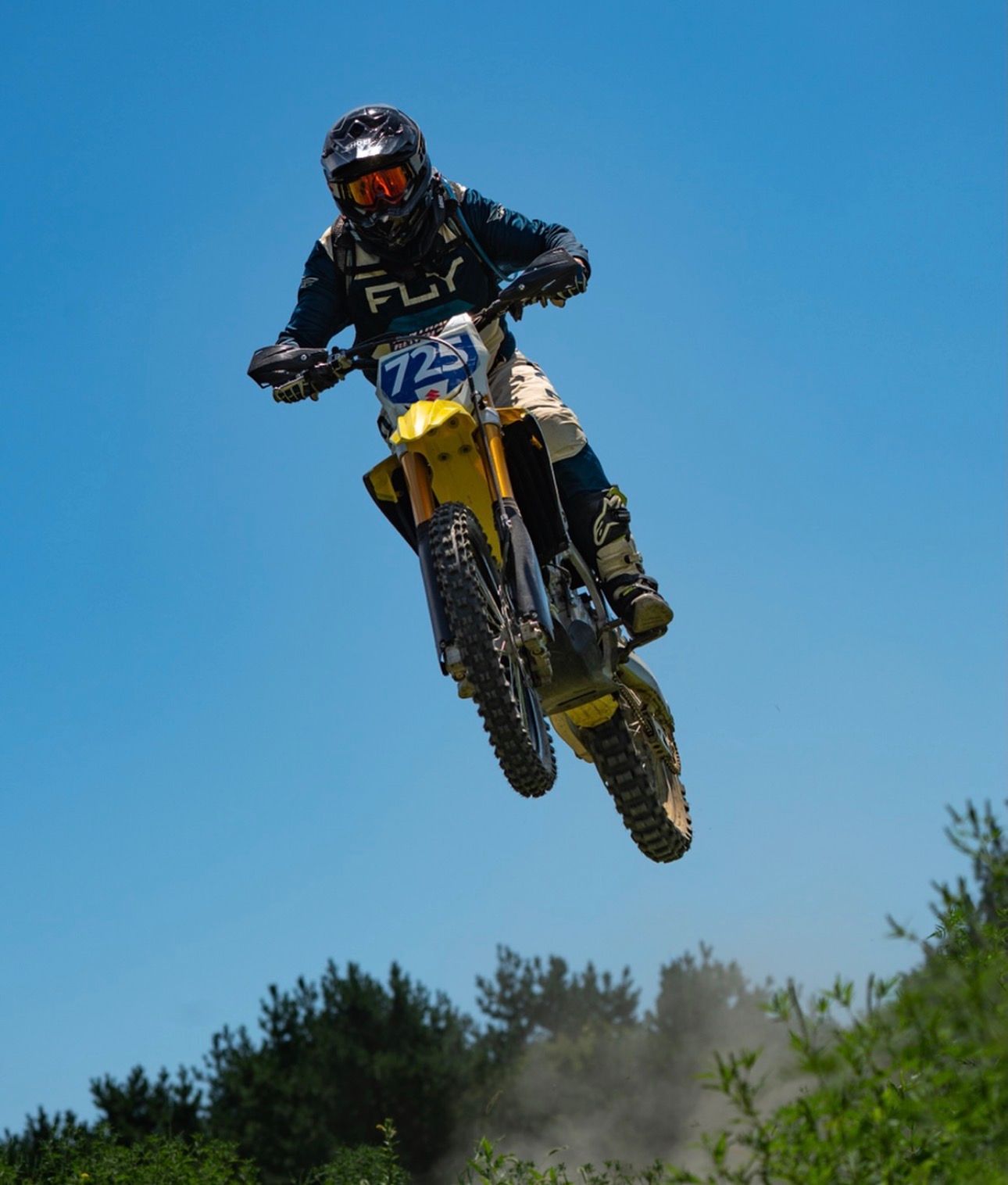 Motocross rider performs aerial jump on yellow dirt bike against bright blue sky during off-road competition.