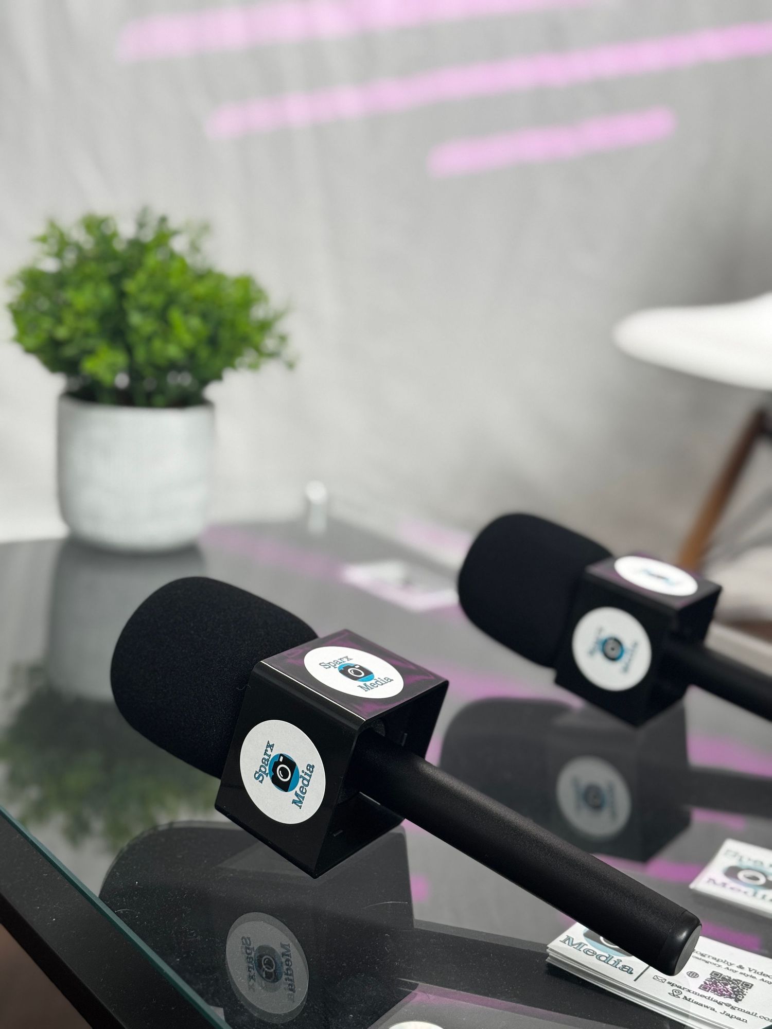 Black branded microphones on a glass table in a modern podcast studio with a green plant and white chair in the background.