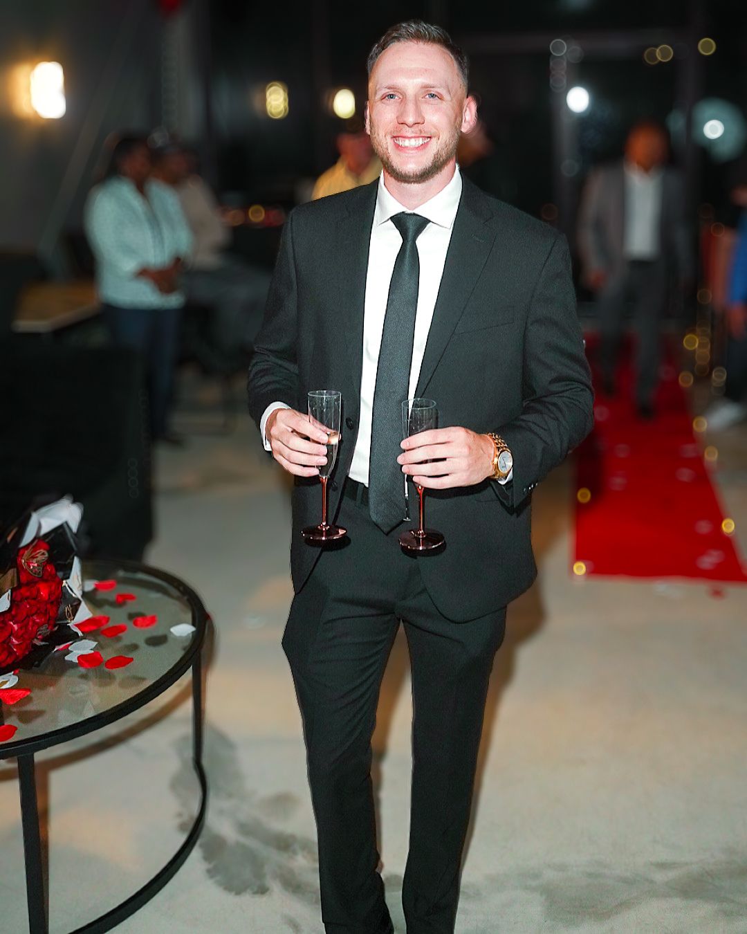 Man in dark suit holding champagne glasses at formal event with red decor.