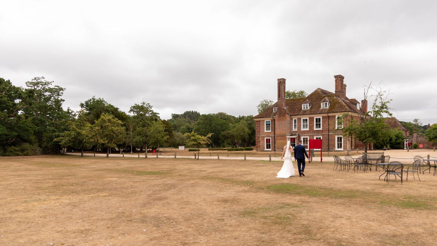 Bride and groom walking hand in hand across a vast lawn beside a grand red brick country house wedding venue.