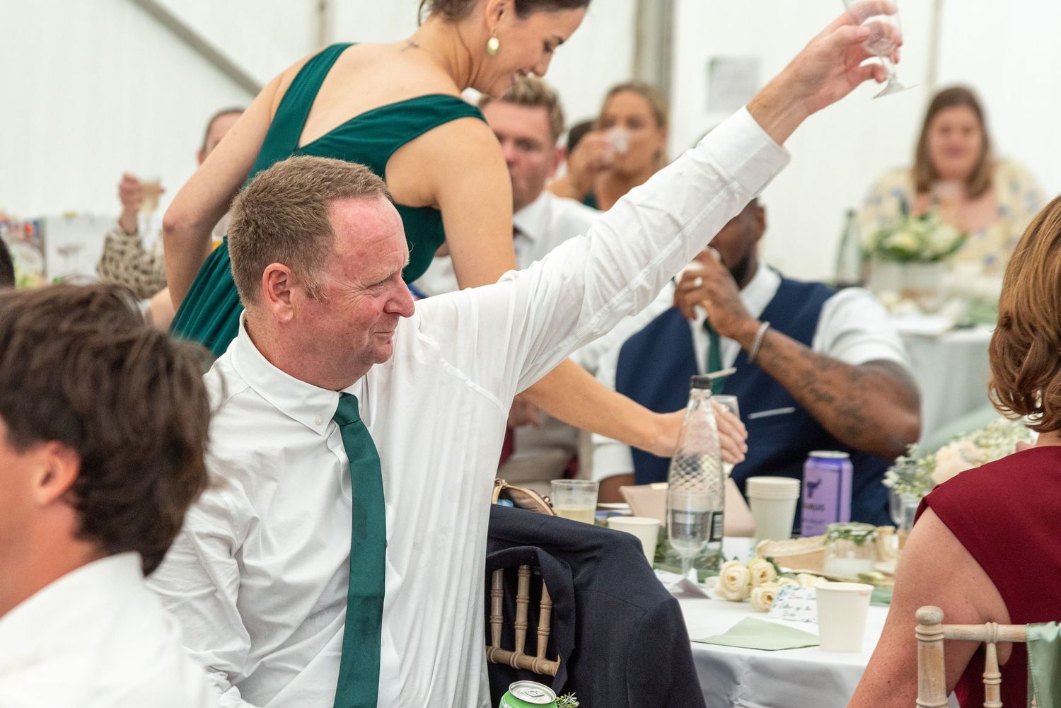 Man in white shirt and green tie raises glass at a wedding reception celebration indoors.